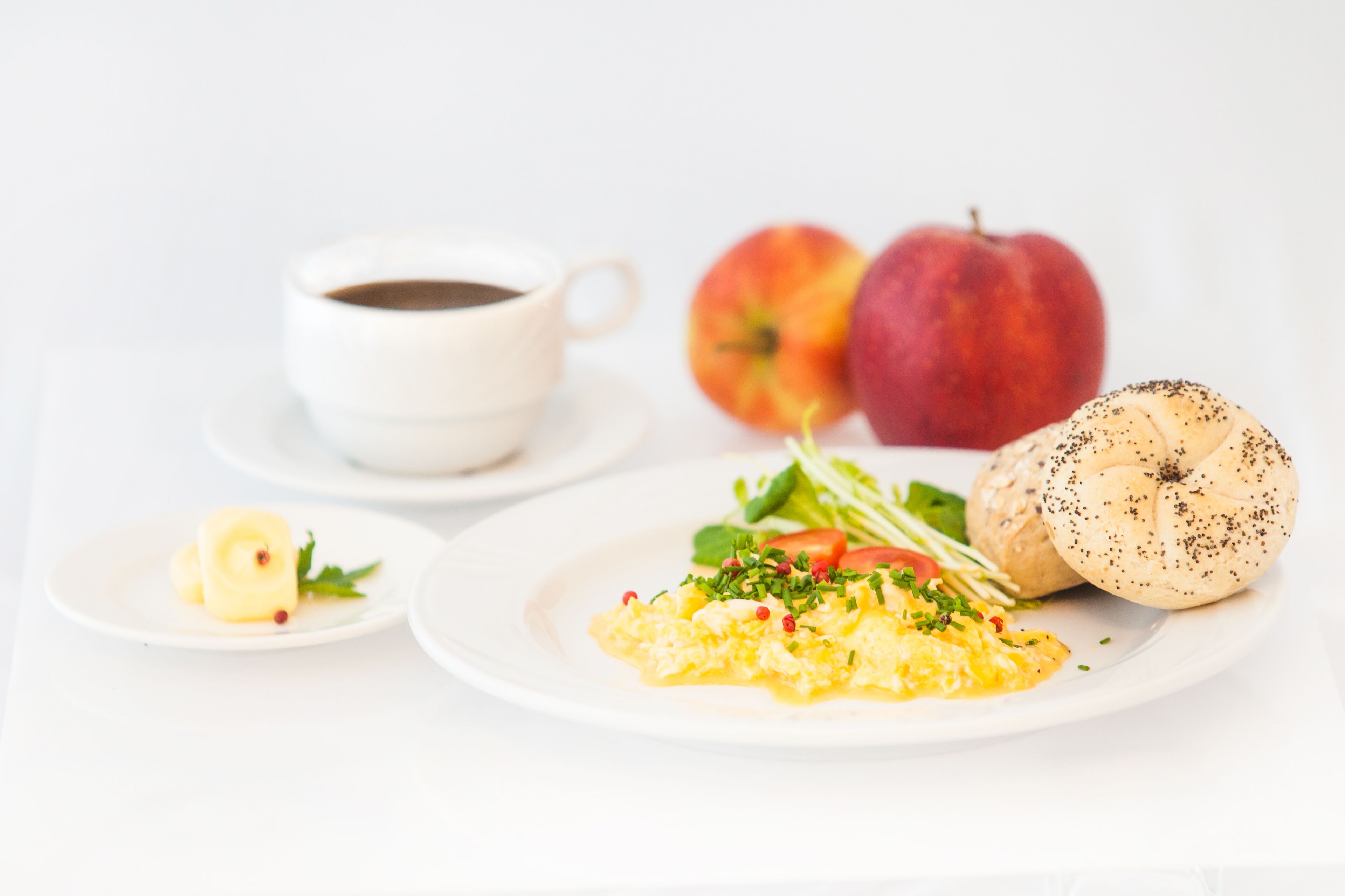 A breakfast setting with scrambled eggs, a croissant, a cup of coffee, and two apples on a white table.