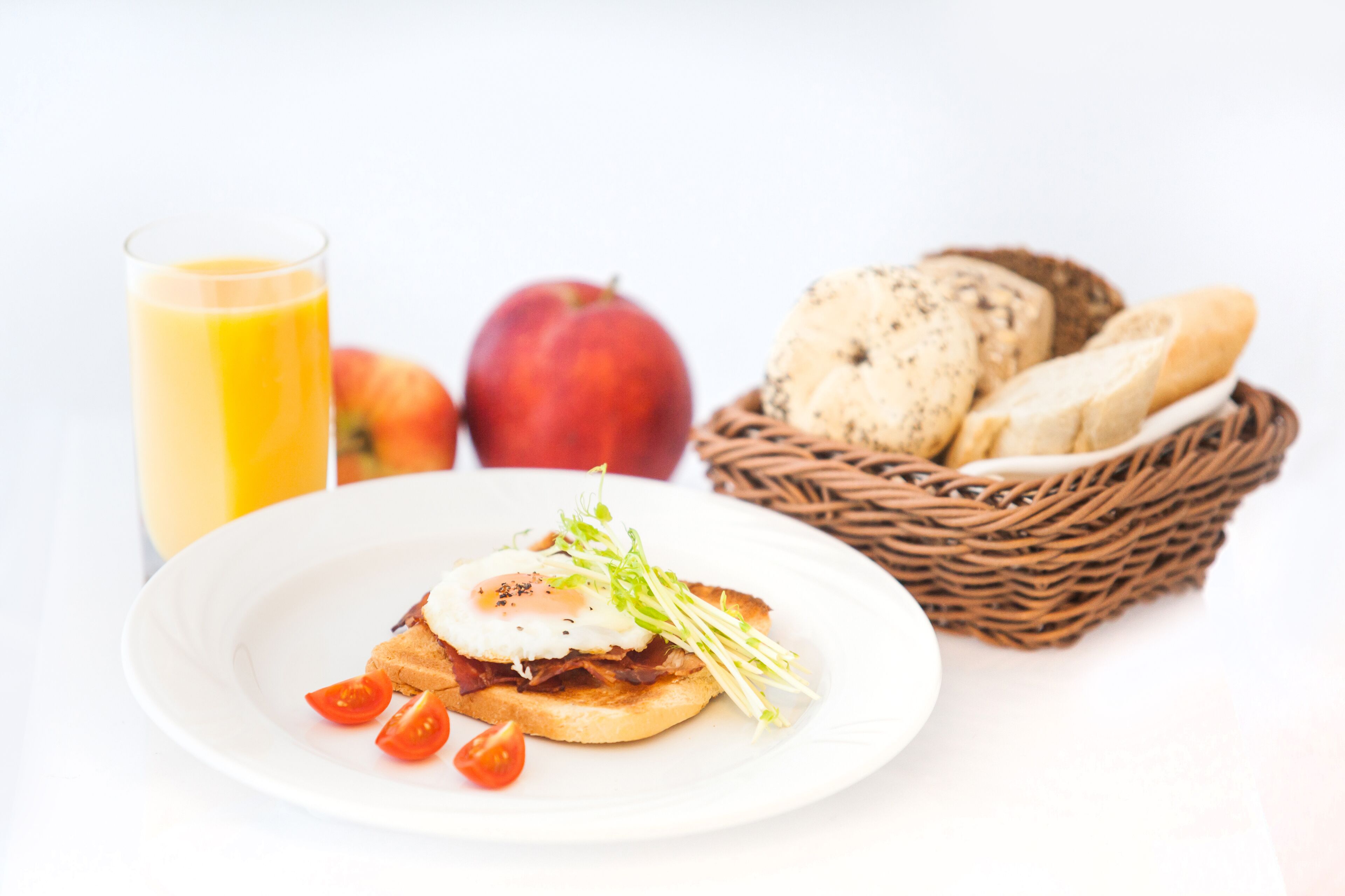 A breakfast plate with toast, egg, and tomato, a glass of orange juice, an apple, and a basket of bread.
