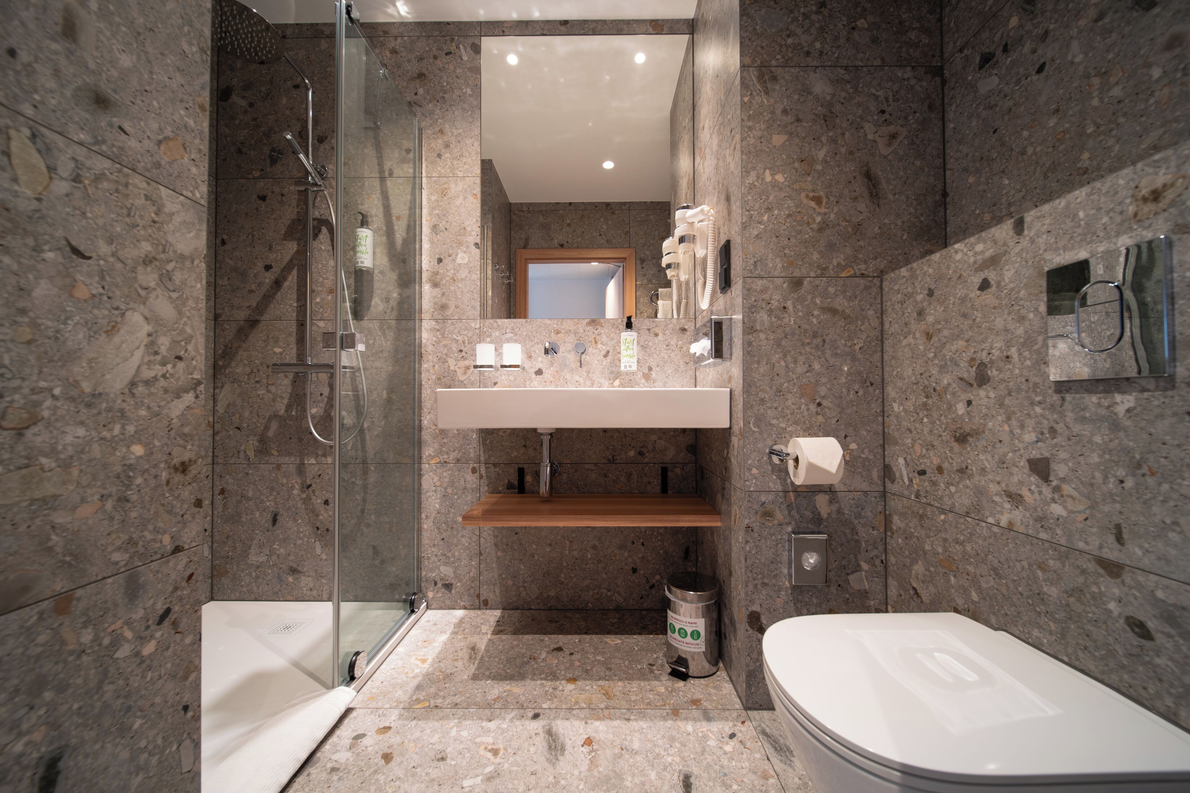 A modern bathroom with a glass shower enclosure, a toilet, a sink with a mirror above, and a trash bin.