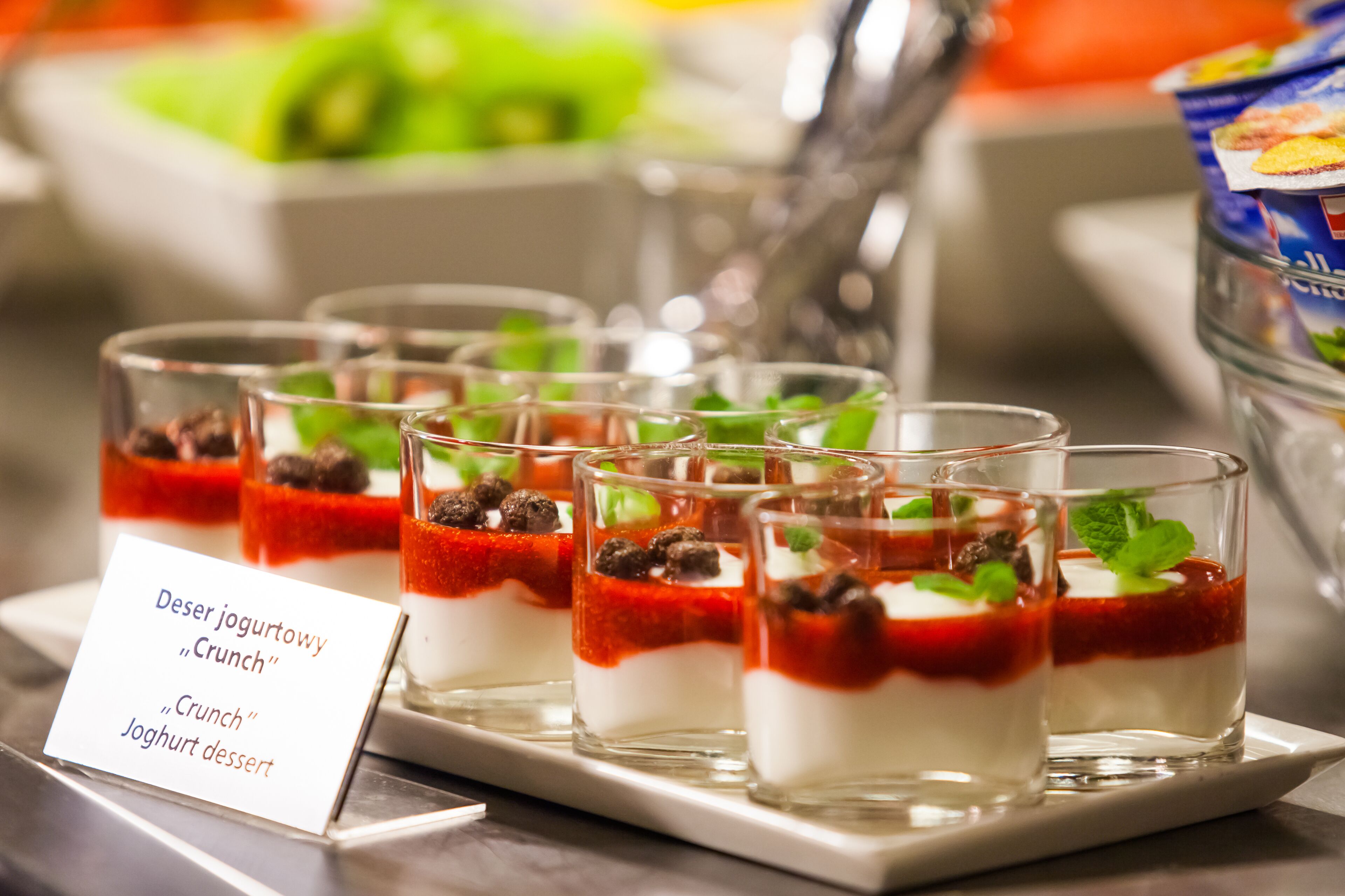 Una estación de postres con vasos para parfait, etiquetados como 'Deser yogurtowy "Crunch"'.