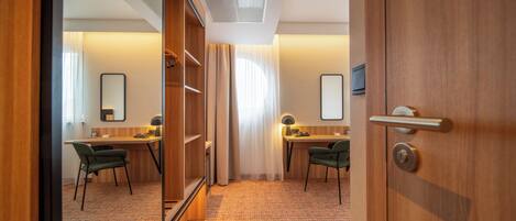 A hotel room with a wooden wardrobe, a desk with a chair, a mirror, and a carpeted floor.