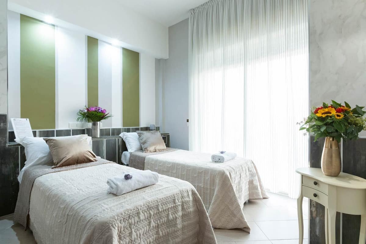 A hotel room with two beds, a large window with sheer curtains, a vase with flowers, and a small table with a vase.