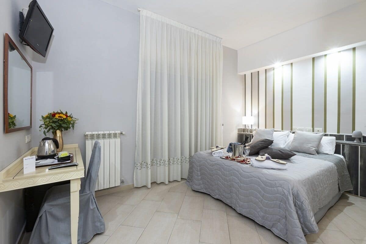 A hotel room with a bed, bedside table, chair, TV, curtains, and a radiator.