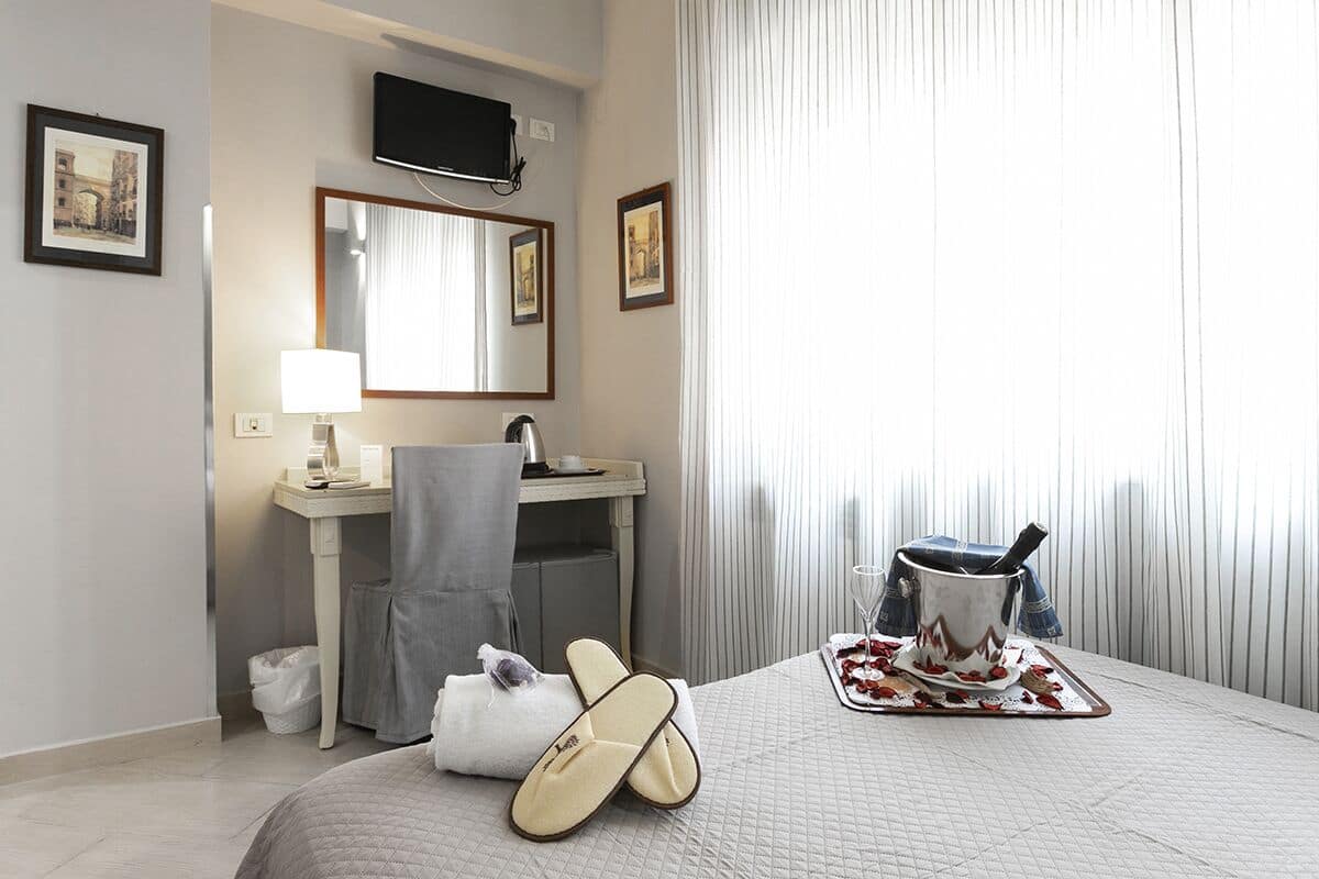 A hotel room with a bed, a bedside table with a lamp, a mirror, a TV mounted on the wall, and a window with curtains.
