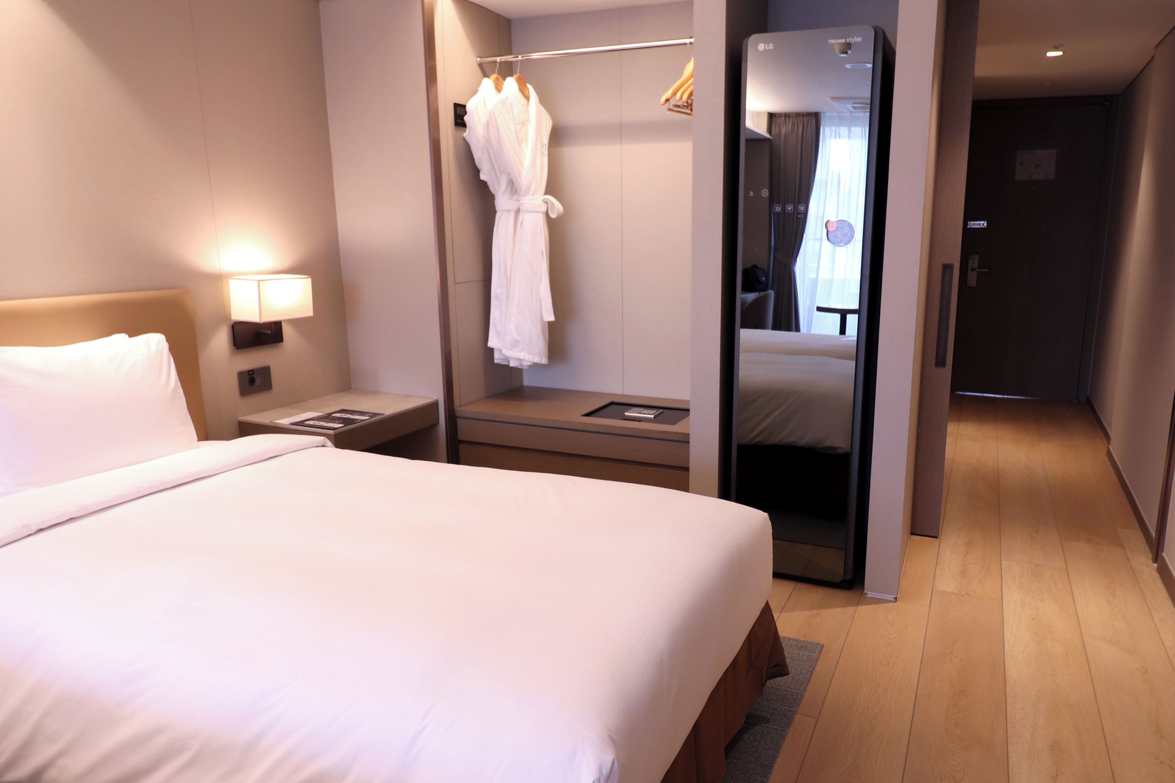 Une chambre d’hôtel avec un grand lit, une table de chevet avec une lampe, un peignoir suspendu à un porte-manteau et un miroir qui reflète la pièce.