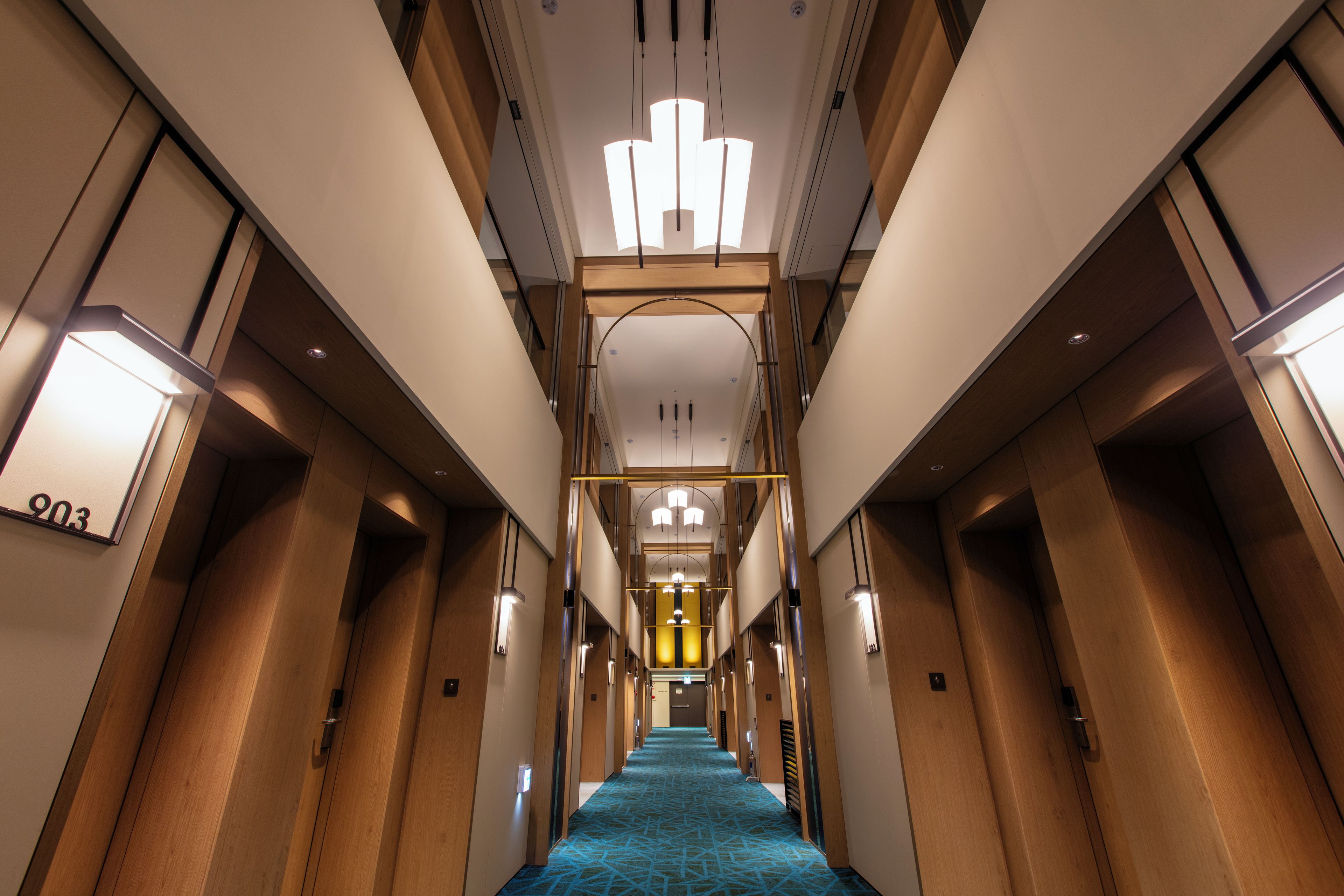 A hotel hallway with a blue carpet, wooden doors, and a central light fixture.