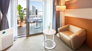 A hotel room with a sofa, a small table with a vase, a lamp, and a view of a cityscape through a glass door.