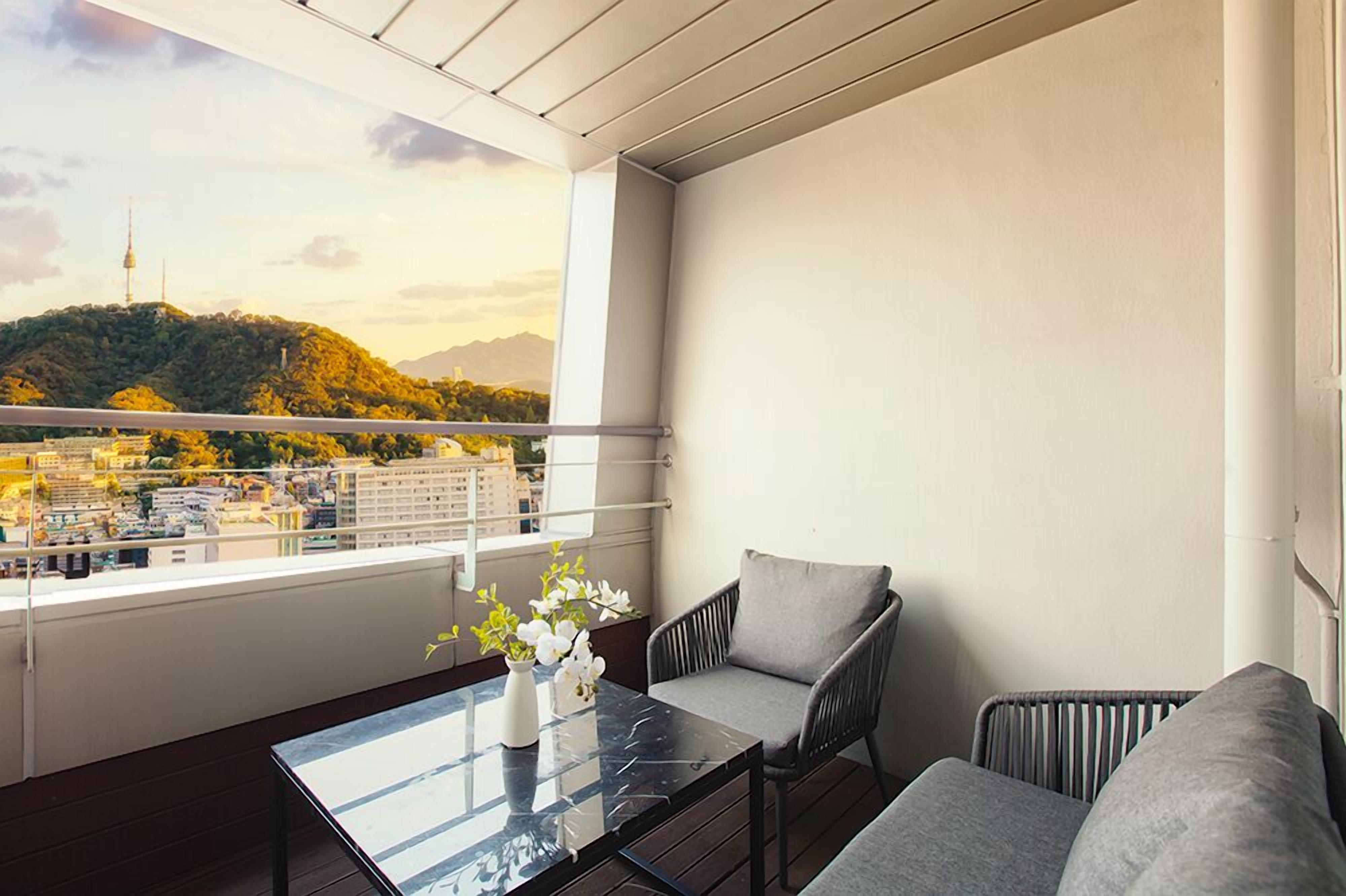 A balcony with a view of a cityscape and mountains, a glass table with a vase of flowers, and two grey chairs.