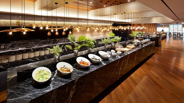 A modern hotel lobby with a long marble counter displaying various food items, pendant lighting, and a wooden floor.