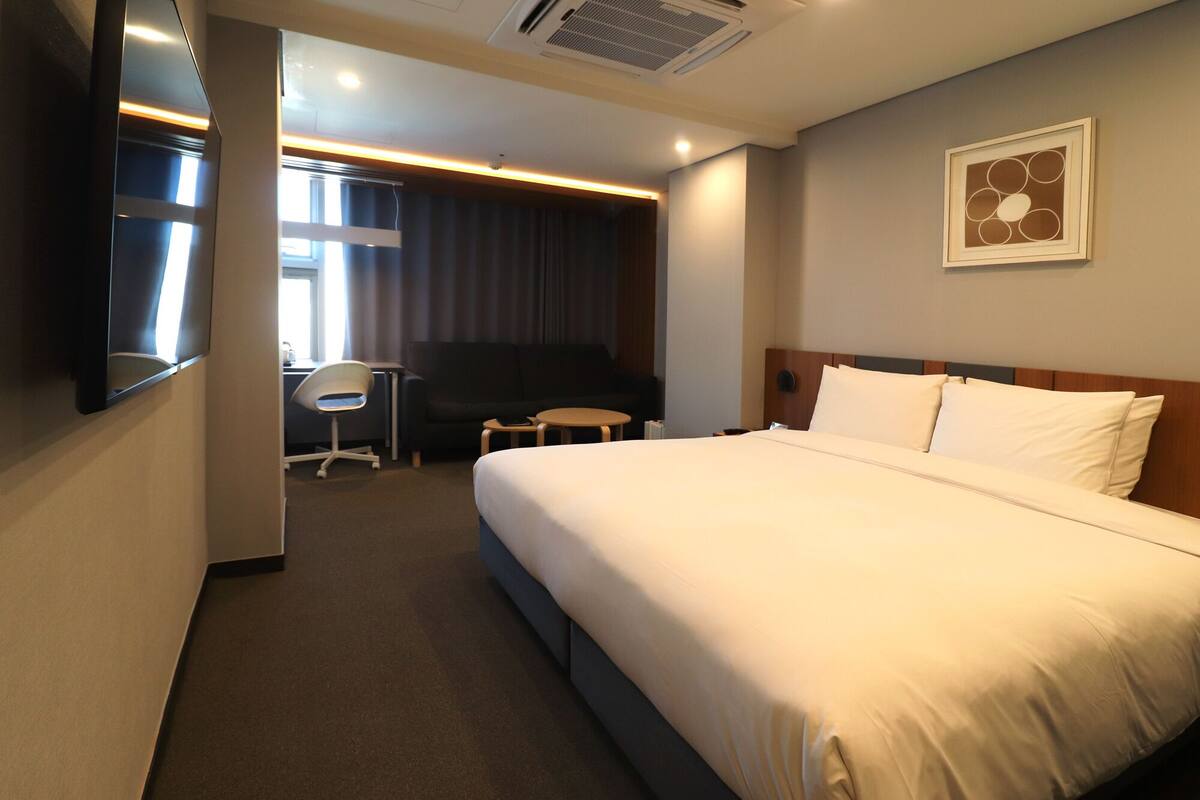 A hotel room with a large bed, a desk with a chair, a small round table, and a TV mounted on the wall.