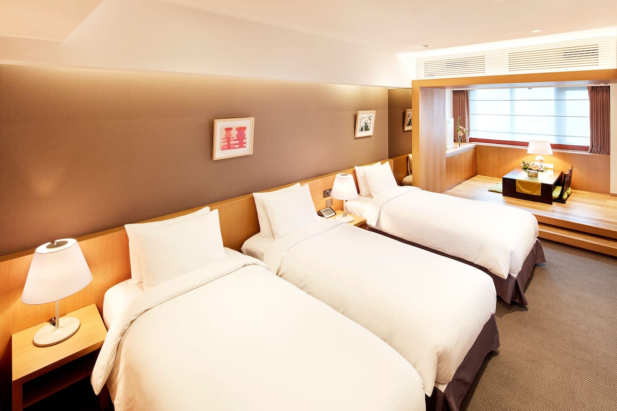 A hotel room with two beds, a nightstand with a lamp, a small table with a lamp, and a window with curtains.