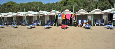 A row of beach huts with blue lounge chairs in front, a red table, and a person sitting on a chair.