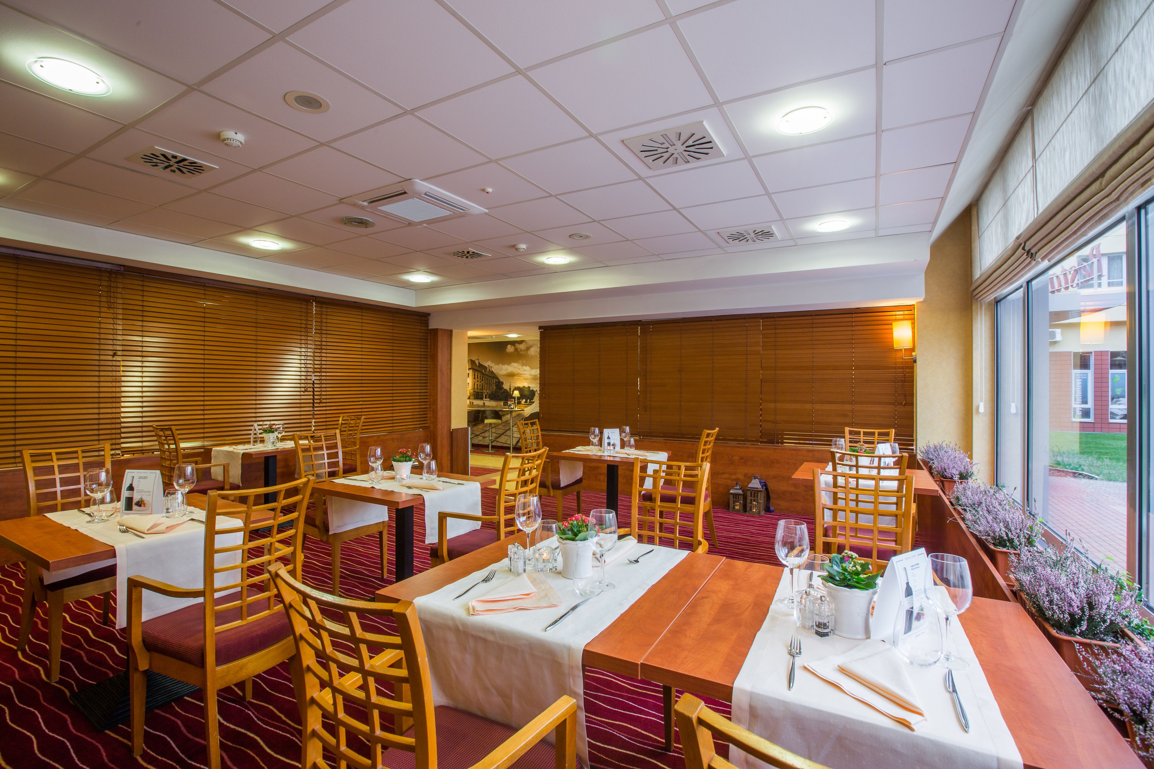 Un restaurant amb diverses taules i caires, una catifa vermella, i vista cap a l'exterior a través de grans finestres.
