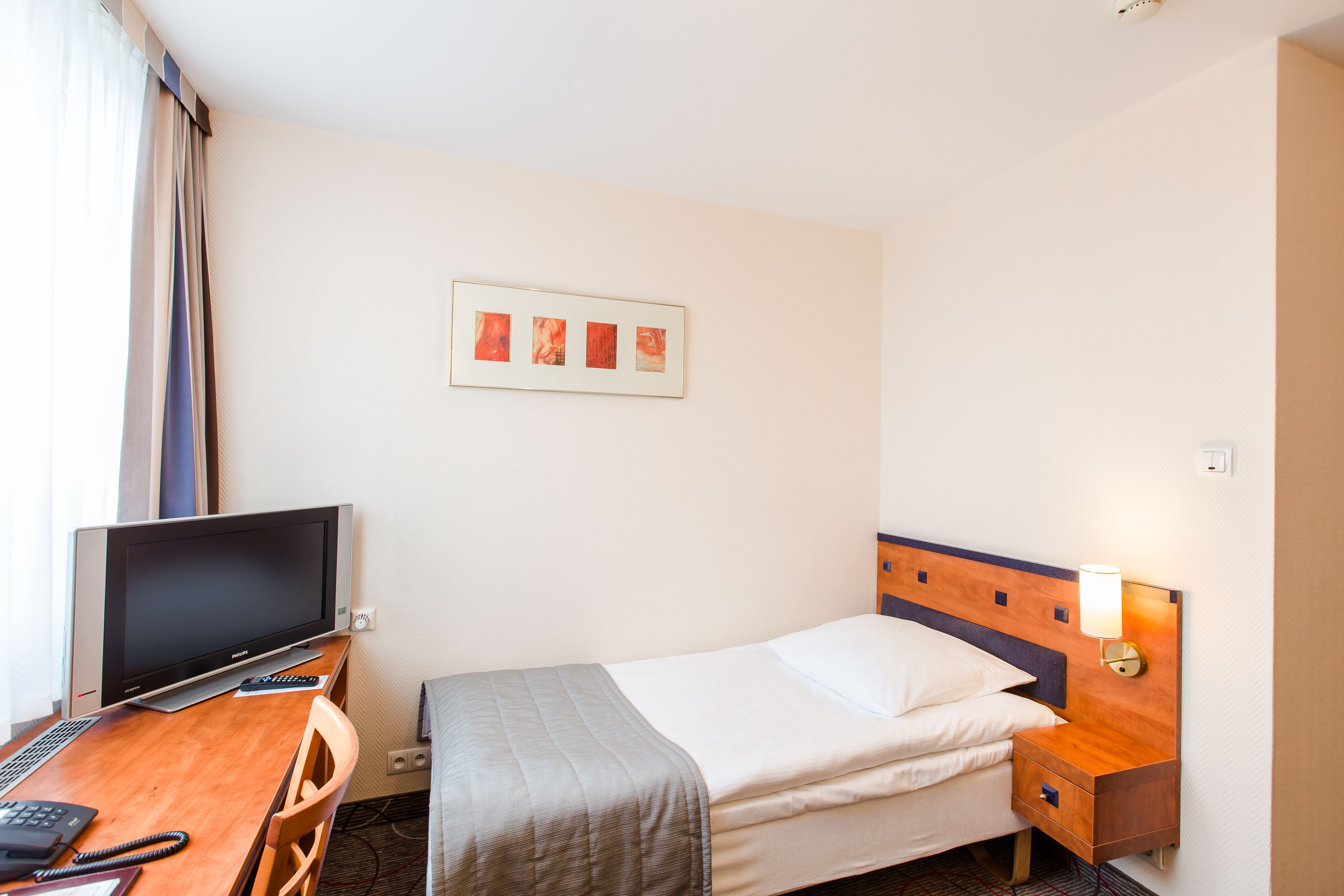 A hotel room with a bed, a desk with a computer, a TV, and a wall-mounted lamp.