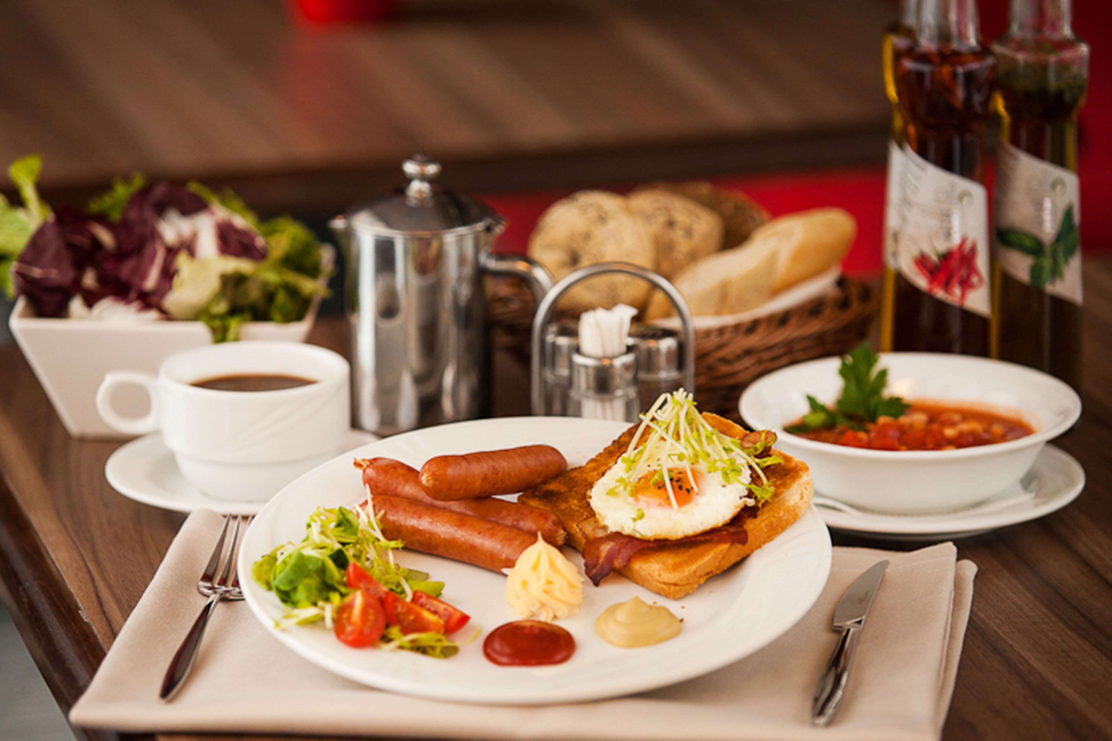 Une table de petit-déjeuner avec une assiette de saucisses, des œufs et des toasts, une tasse de café, un bol de soupe et un panier de pain.