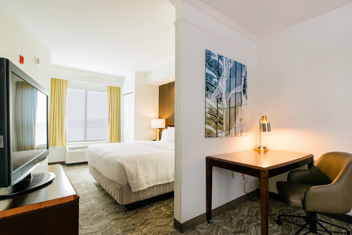 Quarto de hotel com uma cama, uma escrivaninha com abajur, televisão e uma janela grande com cortinas.