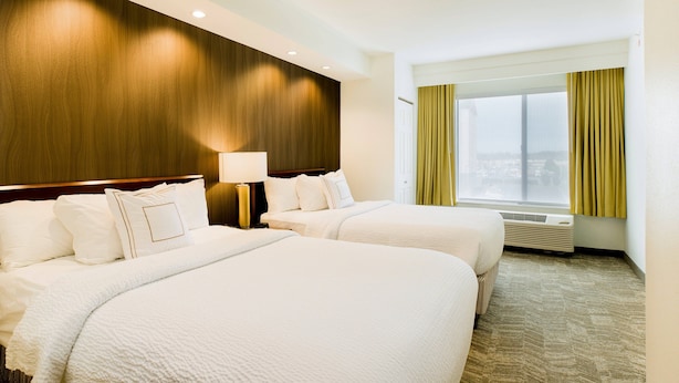 A hotel room with two beds, a wooden headboard, a window with curtains, and a wall-mounted air conditioner.