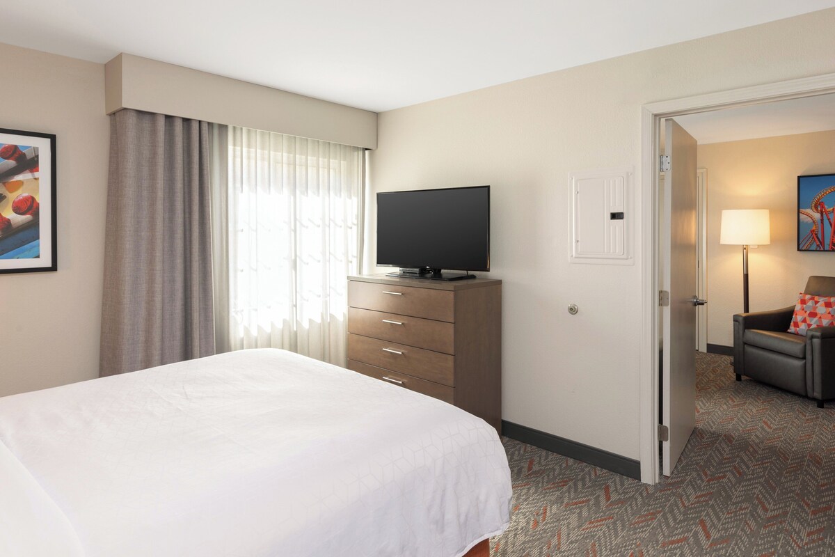 A hotel room with a large bed, a flat-screen TV on a wooden dresser, a door leading to another room, and a lamp on a side table.