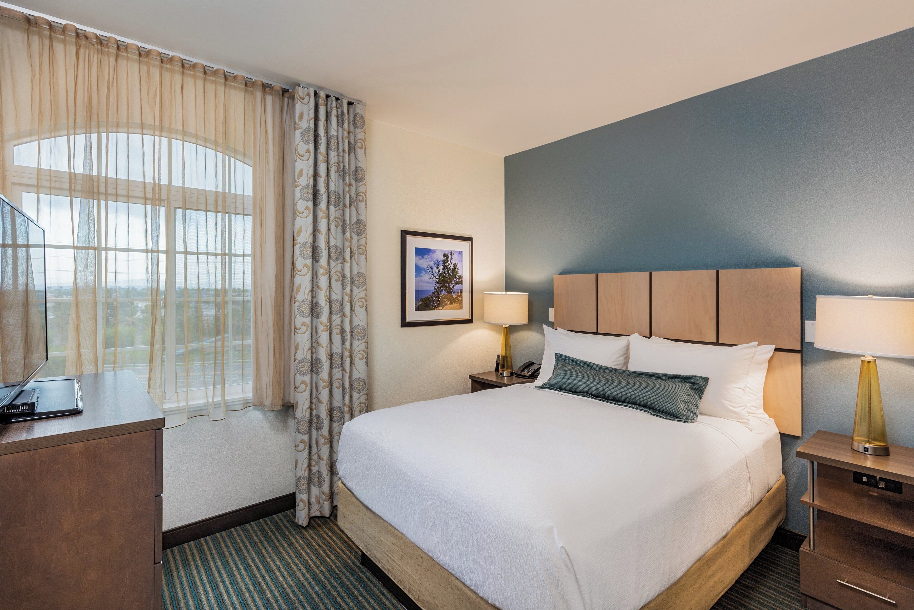 A hotel room with a large bed, a desk, a chair, a nightstand with a lamp, and a framed picture on the wall.