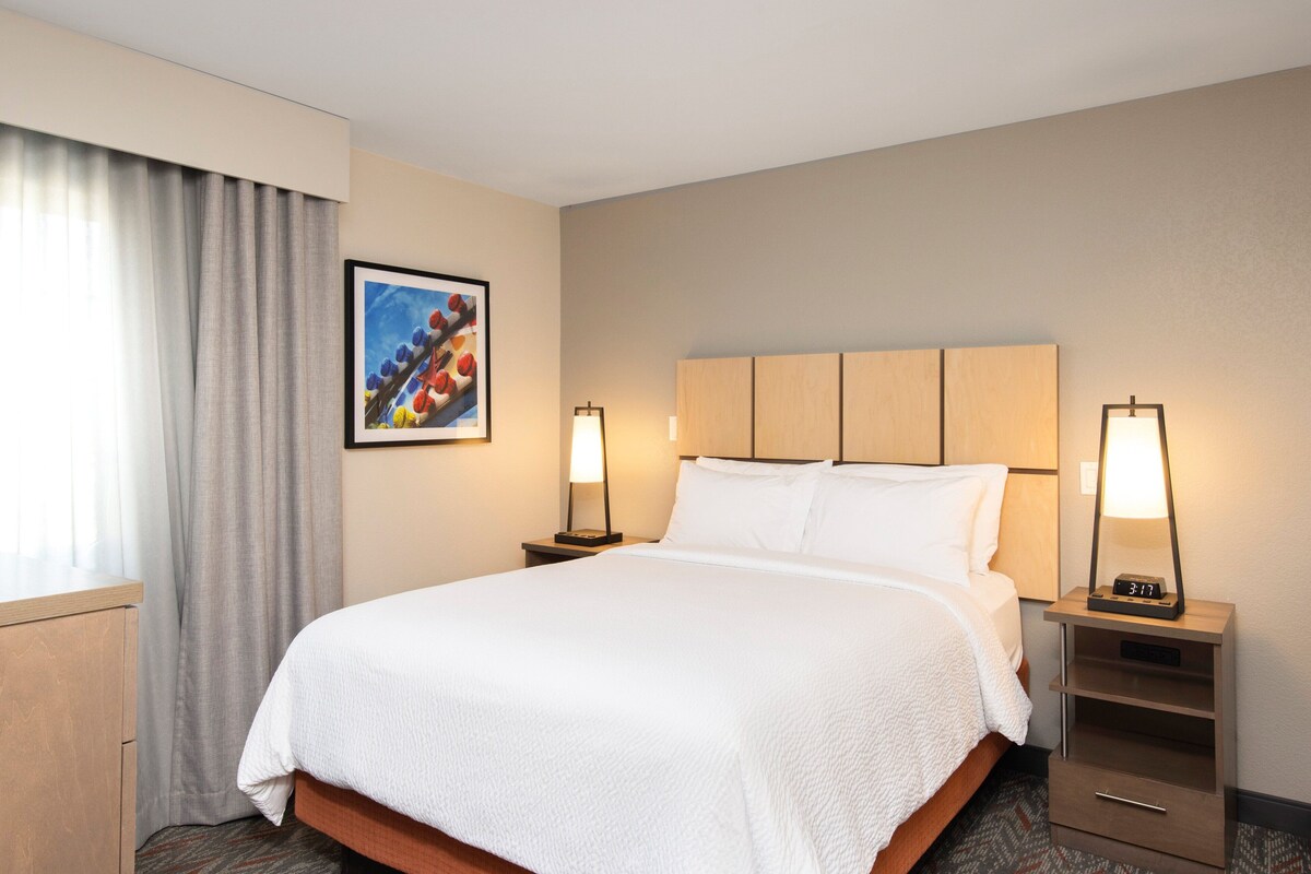A hotel room with a large bed, two bedside tables with lamps, a wooden headboard, a framed picture on the wall, and a window with curtains.