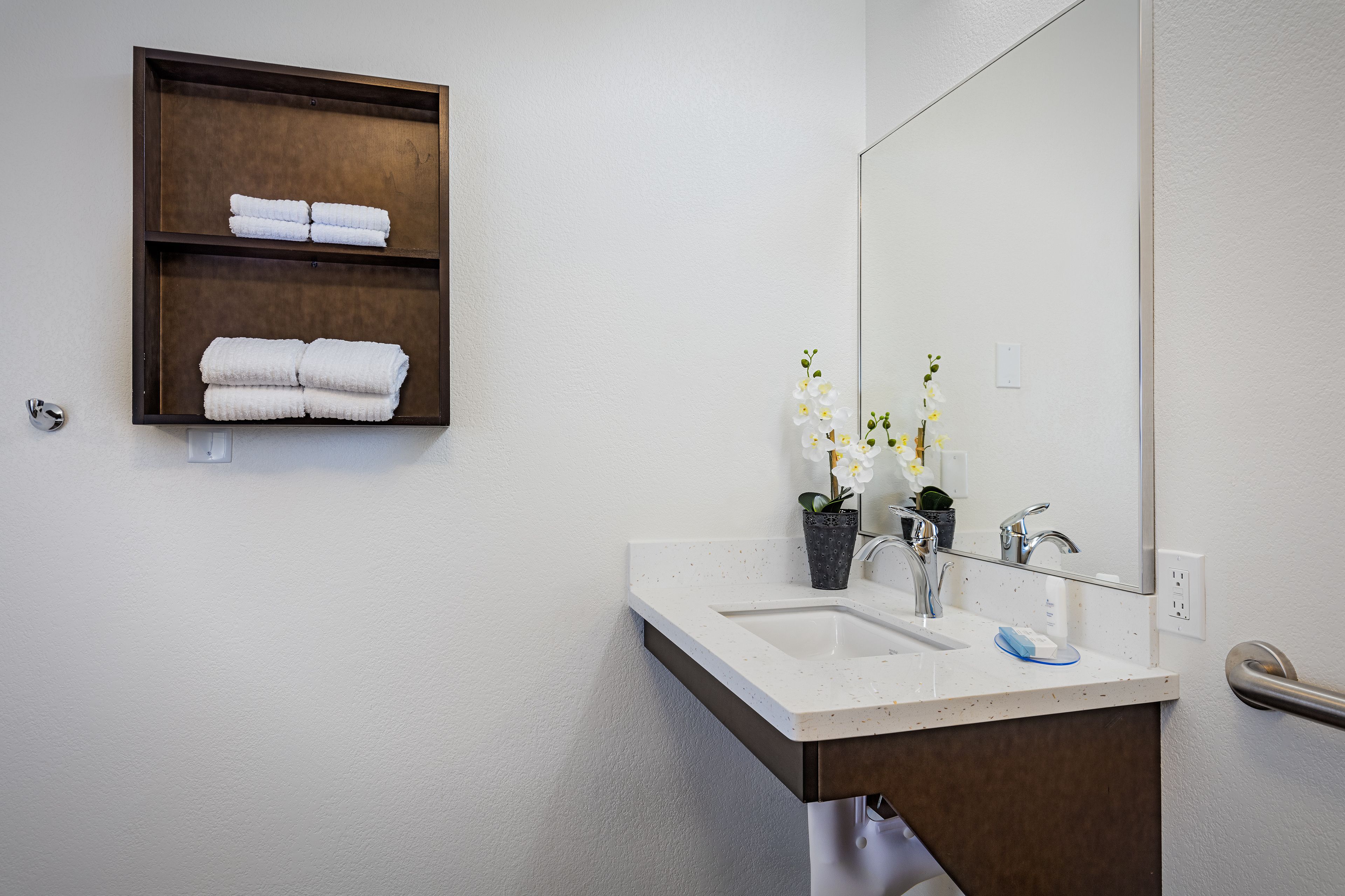 A bathroom with a white sink, two faucets, a mirror, and a wooden shelf with folded towels.