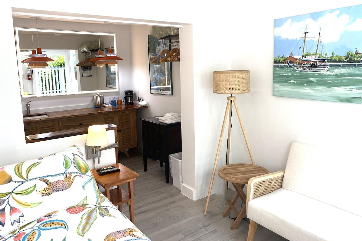 A room with a bed, a desk, a chair, a lamp, a painting of a boat on water, and a view of the sea.