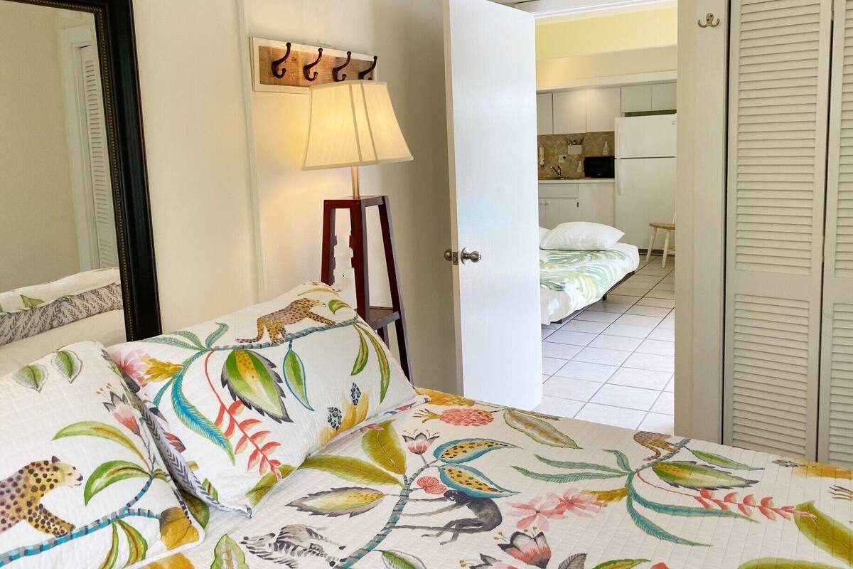 A bedroom with a bed featuring a colorful floral and animal print bedspread, a bedside lamp, and a wooden chair.