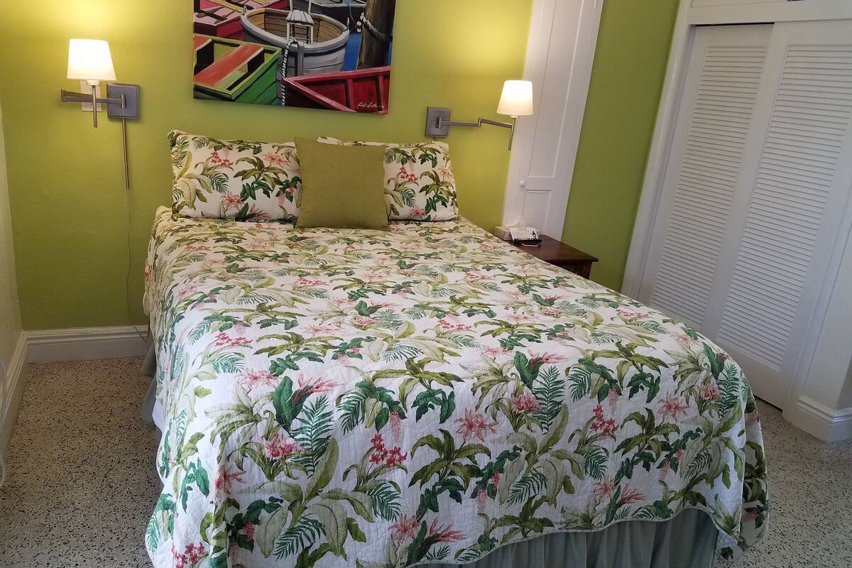 A bed with floral bedding, two wall-mounted lamps, a painting of boats, and a closed door.