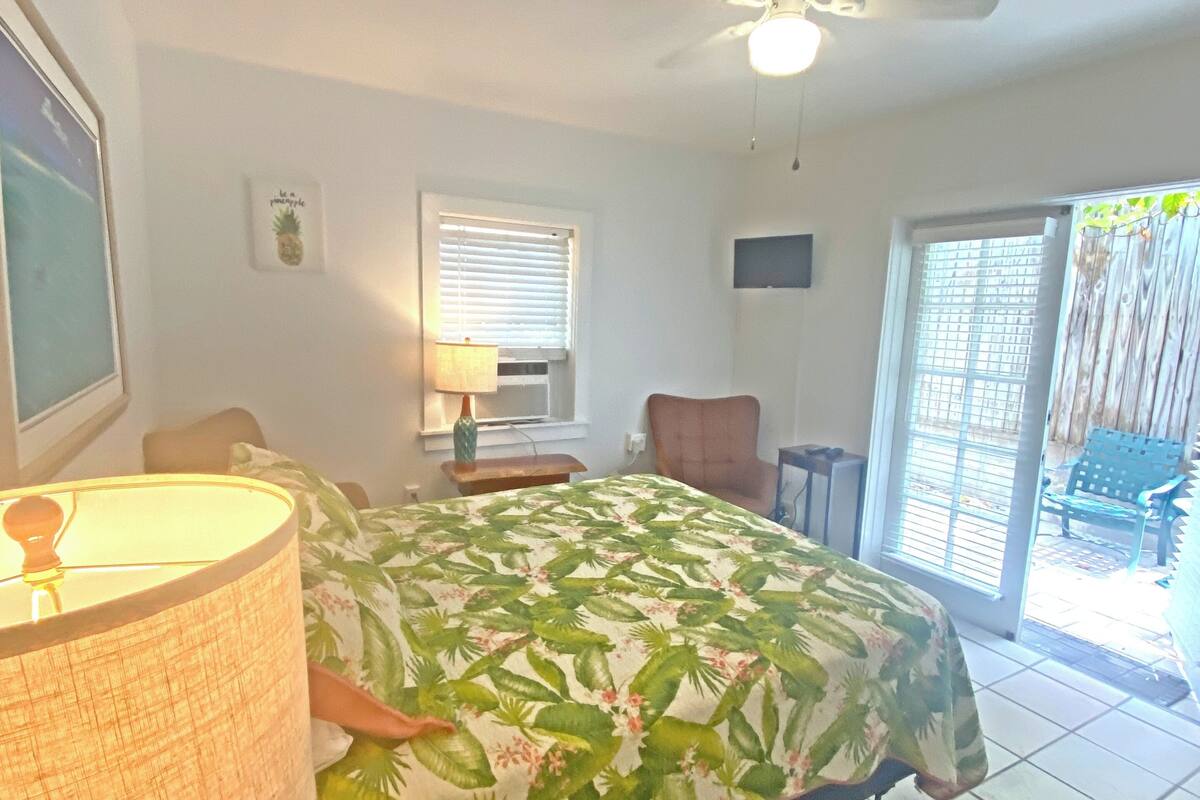 A bedroom with a bed, a ceiling fan, a window with blinds, a chair, and a small table with a lamp.