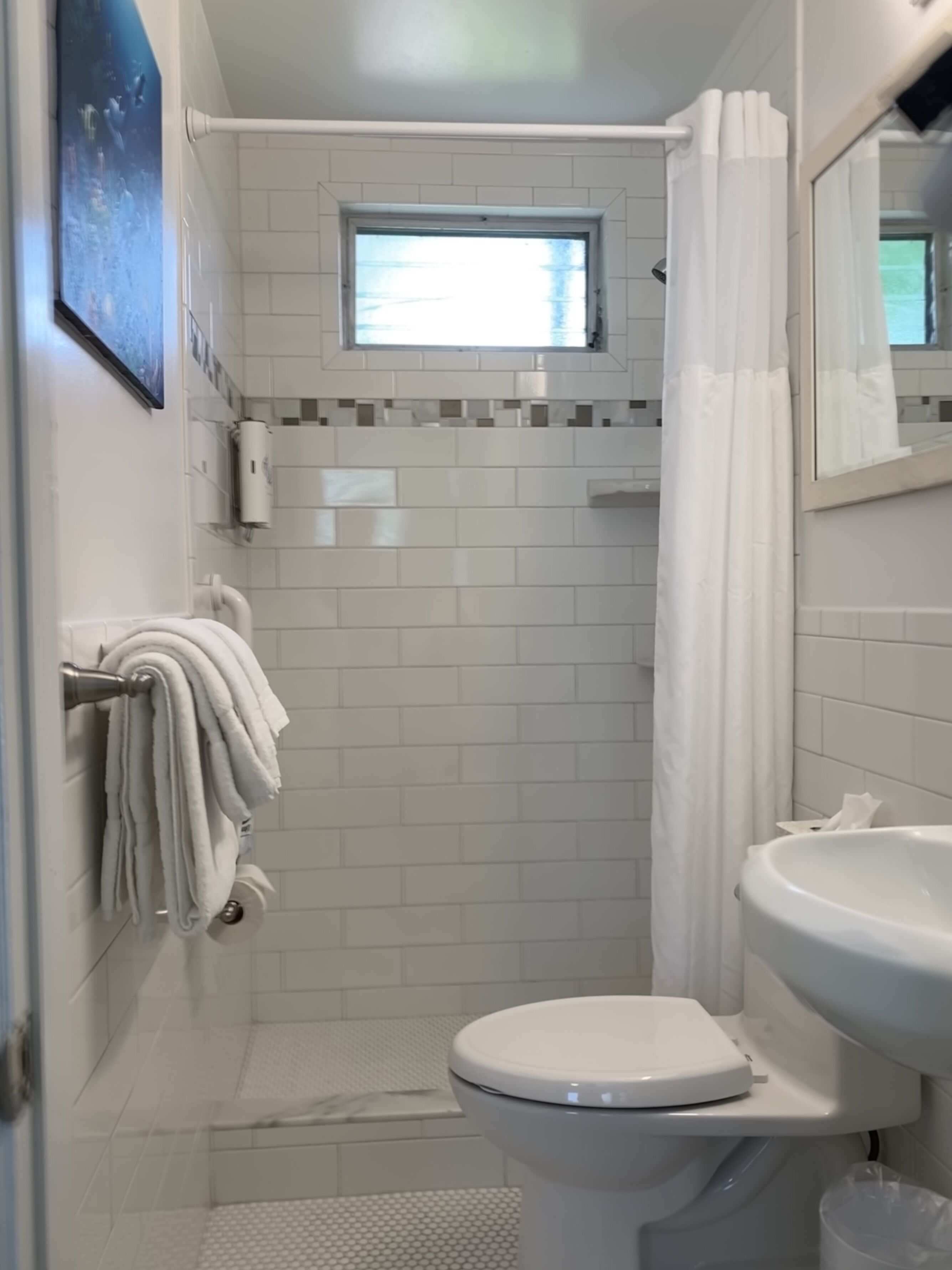 A small, clean bathroom with white tiles, a toilet, a sink, a shower with a curtain, and a window above the shower.