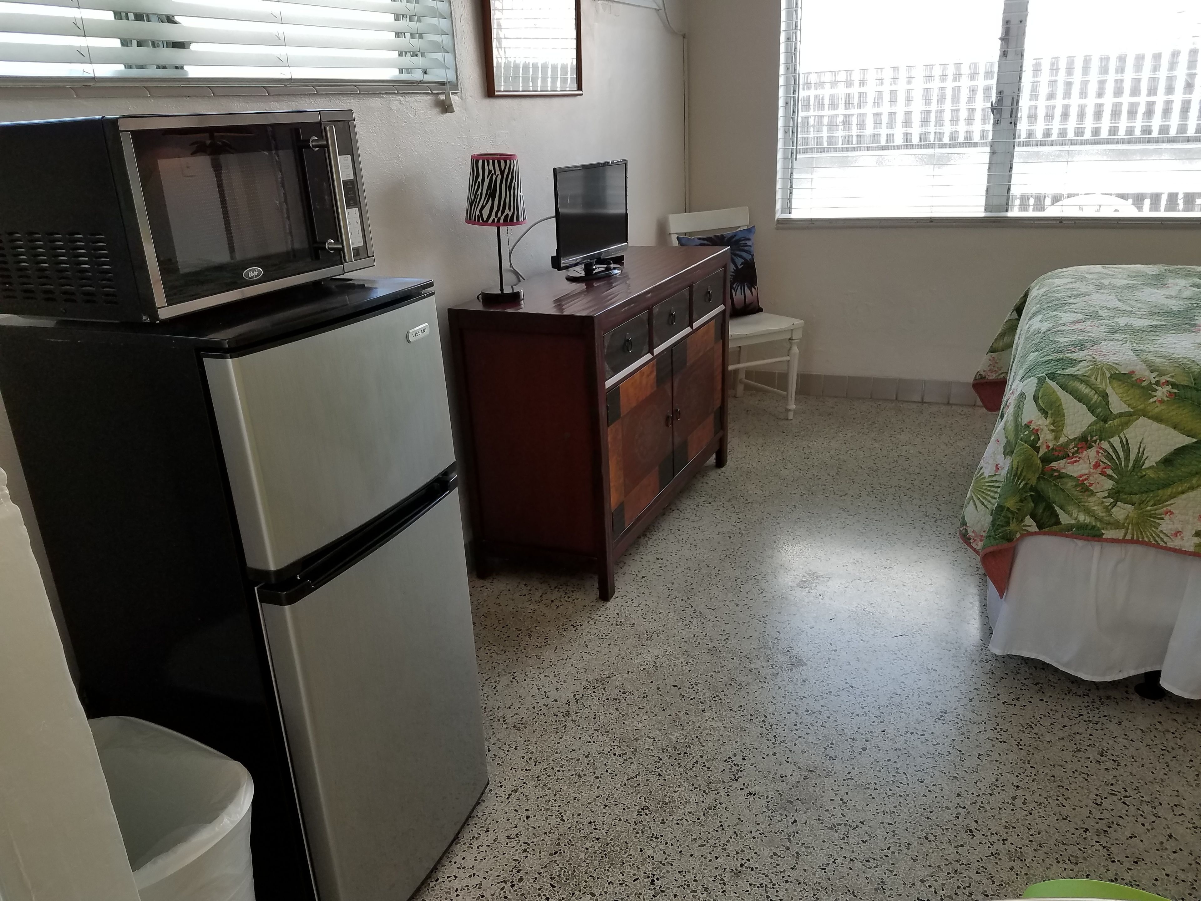 Une chambre compacte dotée d’une kitchenette, d’un lit recouvert d’une couvre-lit à motifs tropicaux, d’une télévision et d’une fenêtre donnant sur des immeubles.