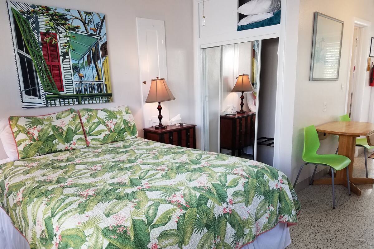 A bedroom with a bed, a nightstand, a table with chairs, a mirror, and a door leading to another room.