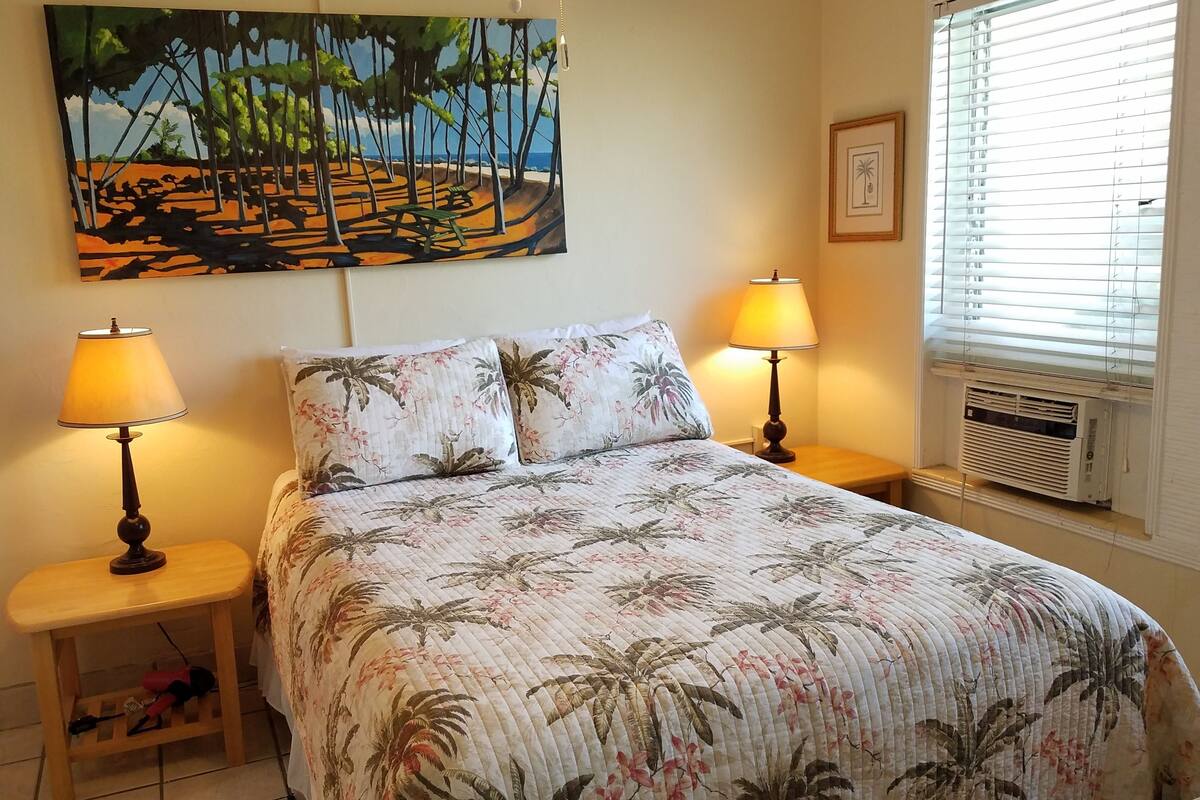 A bedroom with a bed, two bedside lamps, a painting, and a window with blinds.