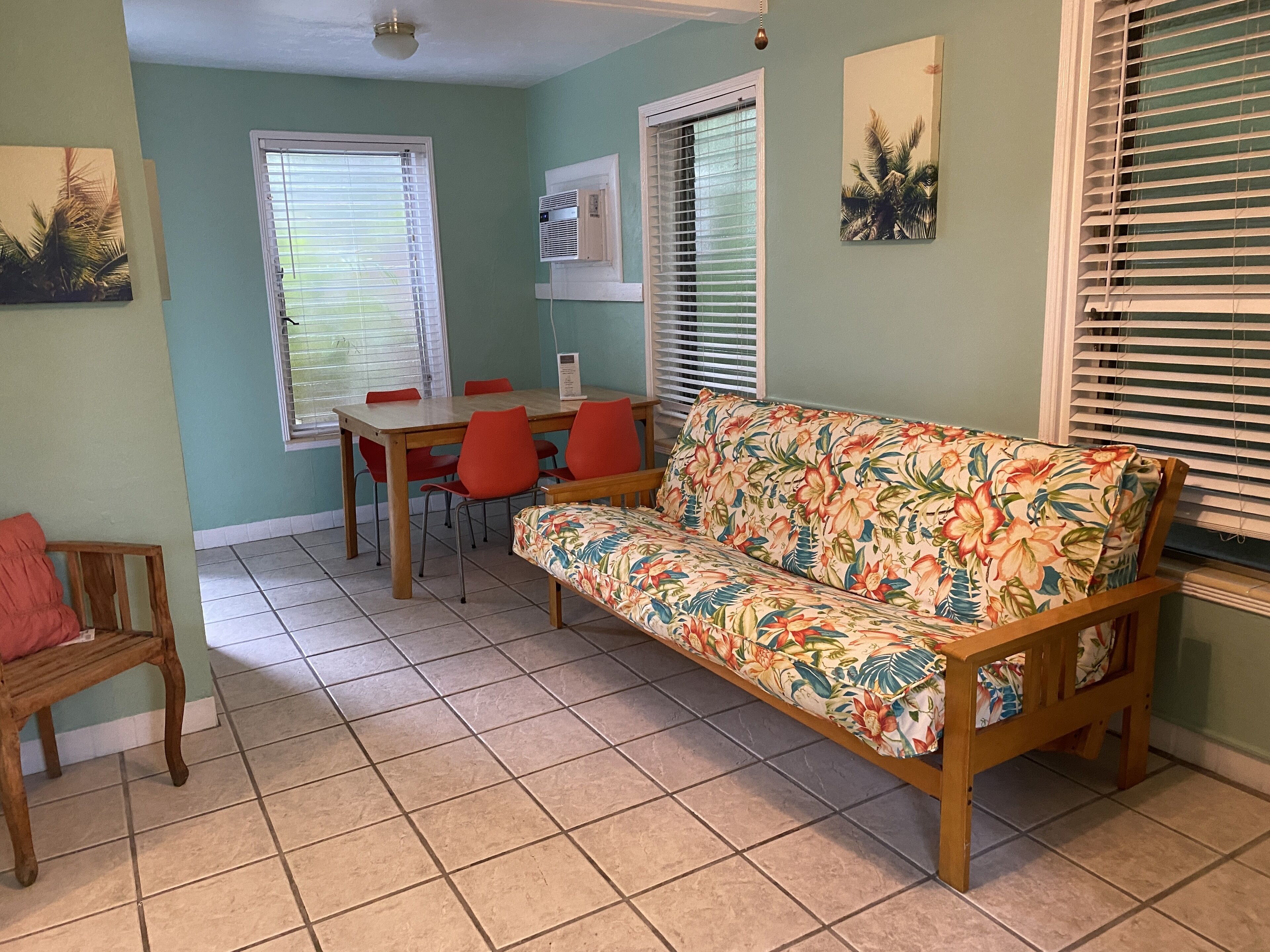 A room with a floral sofa, a dining table with chairs, and a window with blinds.
