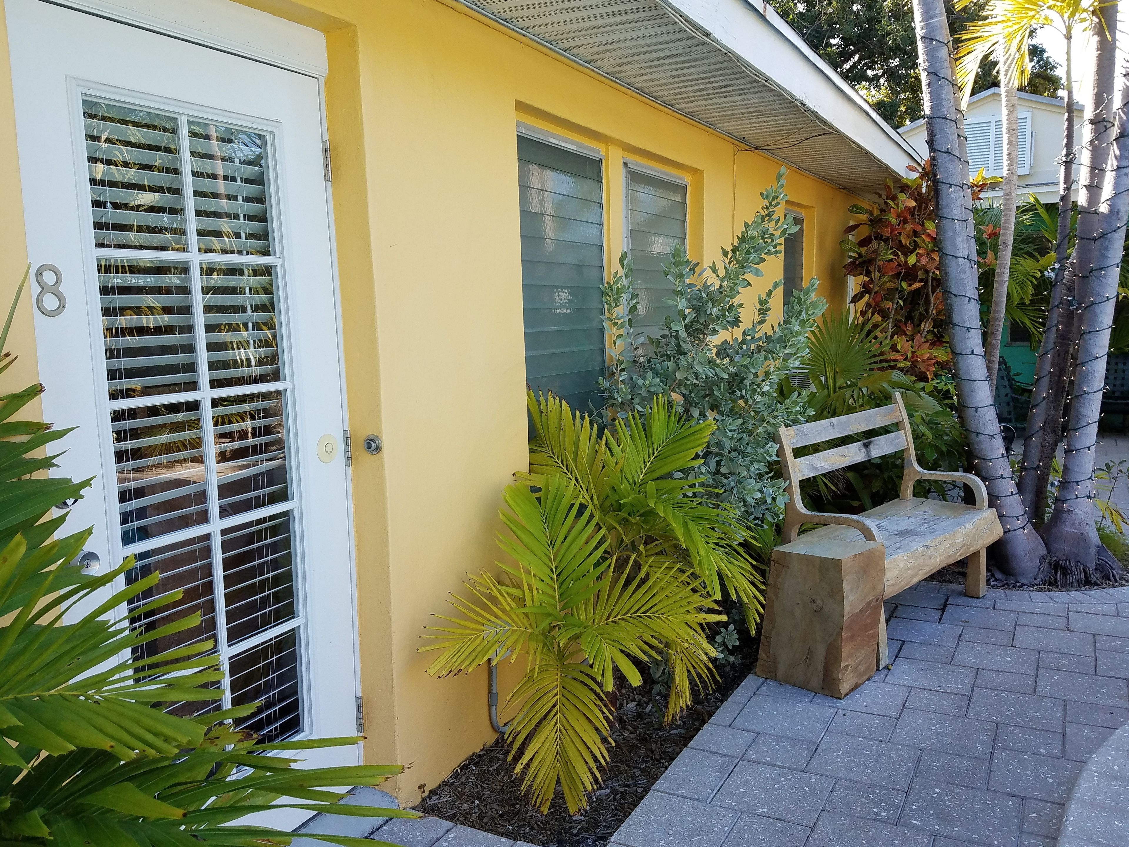 A yellow house with a white door and window shutters, a bench, and various plants.
