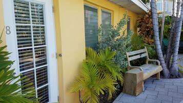 A yellow house with a white door and window shutters, a bench, and various plants.