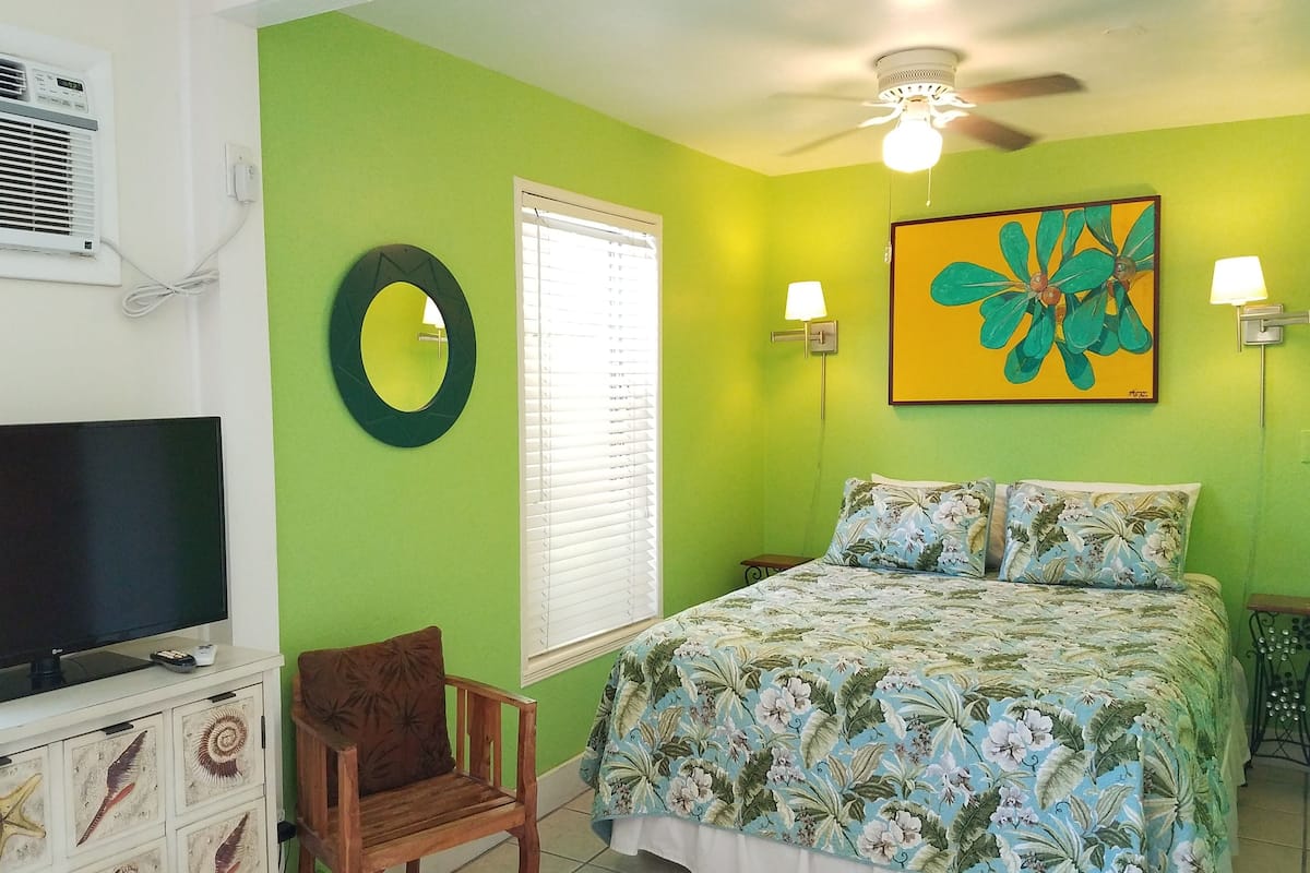 A bedroom with a green wall, a bed with floral bedding, a wooden chair, a TV, and a ceiling fan.