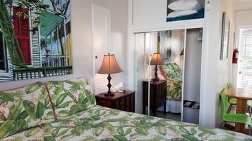 A hotel room with a bed, two bedside tables, a nightstand, a green chair, a table, a painting, a door, and a window with blinds.