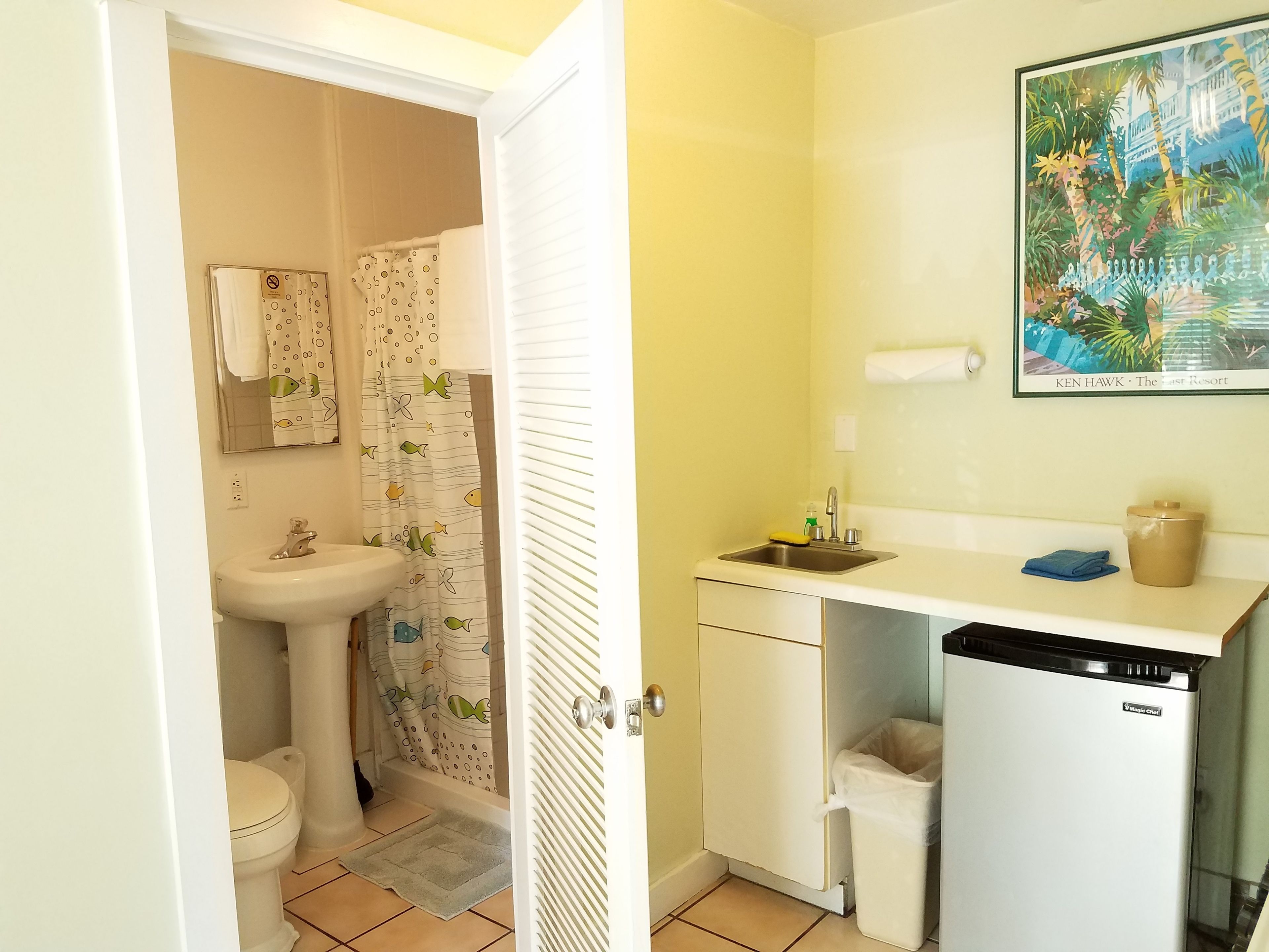 A compact bathroom with a toilet, sink, and shower area. A small refrigerator and a trash bin are visible.