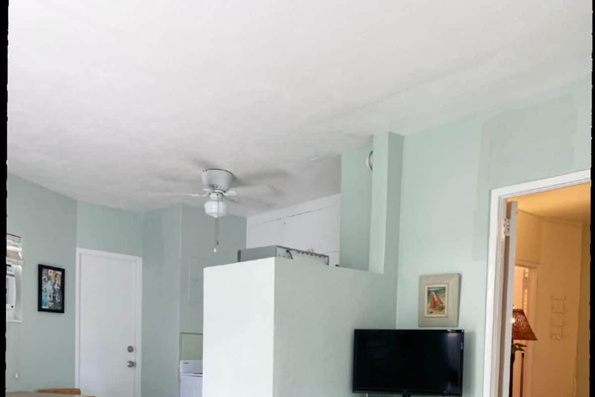 A room with a bed, a television on a stand, a dining table with chairs, and a door leading to another room.