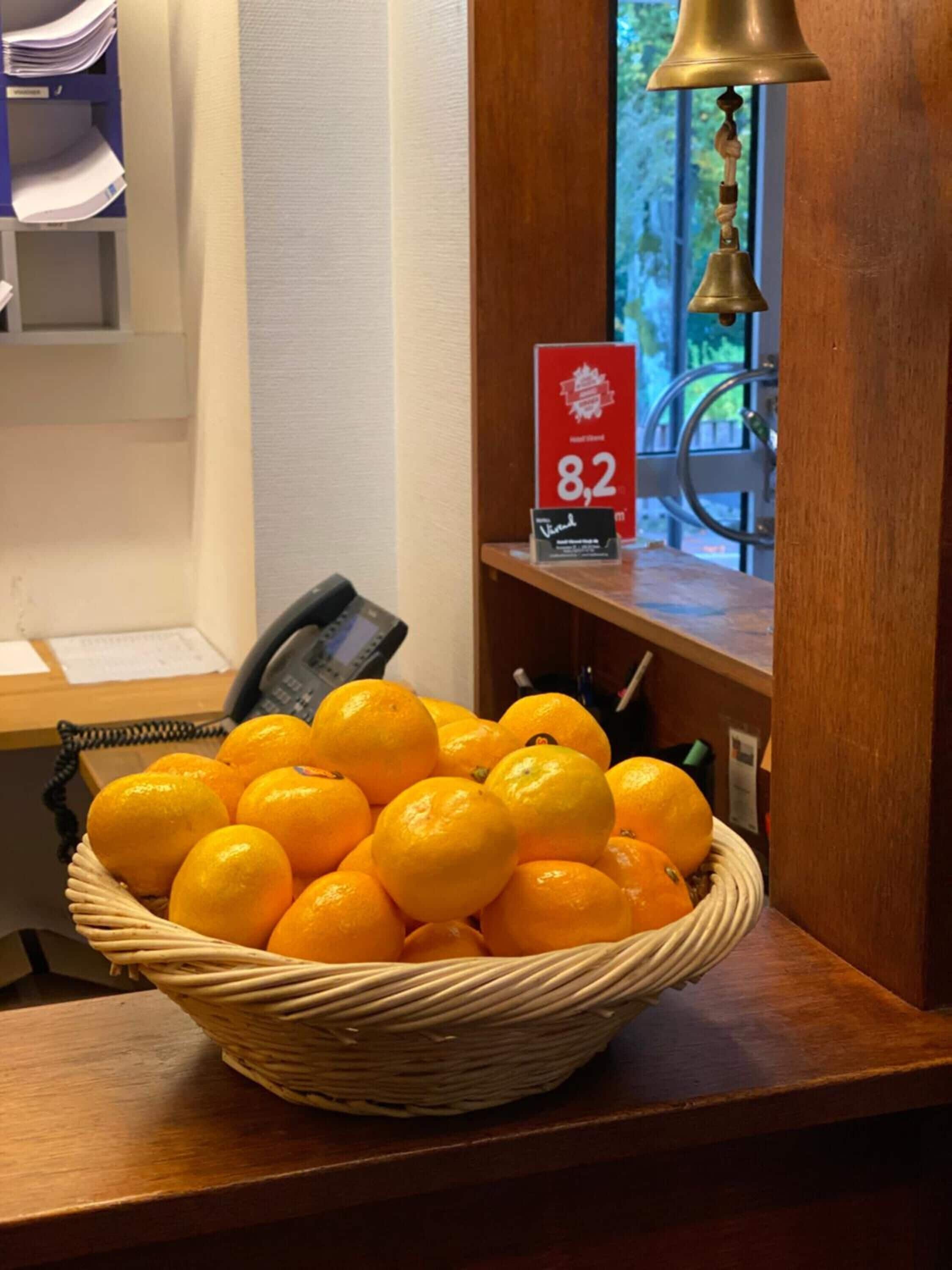 Un panier en osier rempli d’oranges est posé sur un comptoir en bois. On aperçoit, à l’arrière-plan, un panneau rouge portant le chiffre 8,2, un téléphone et une cloche.