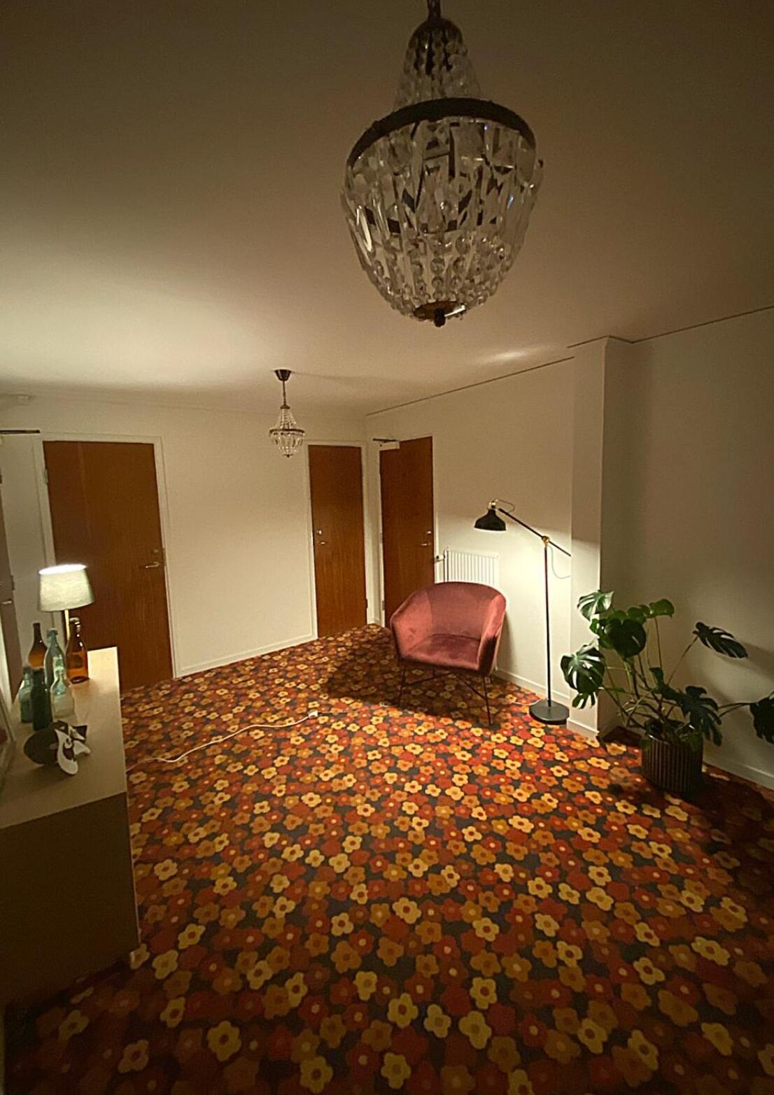 Une pièce avec un tapis à motifs floraux, un lustre, un bureau avec une lampe, une chaise et une plante en pot.