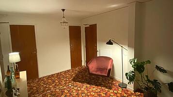 A room with a floral carpet, a chandelier, a desk with a lamp, a chair, and a potted plant.