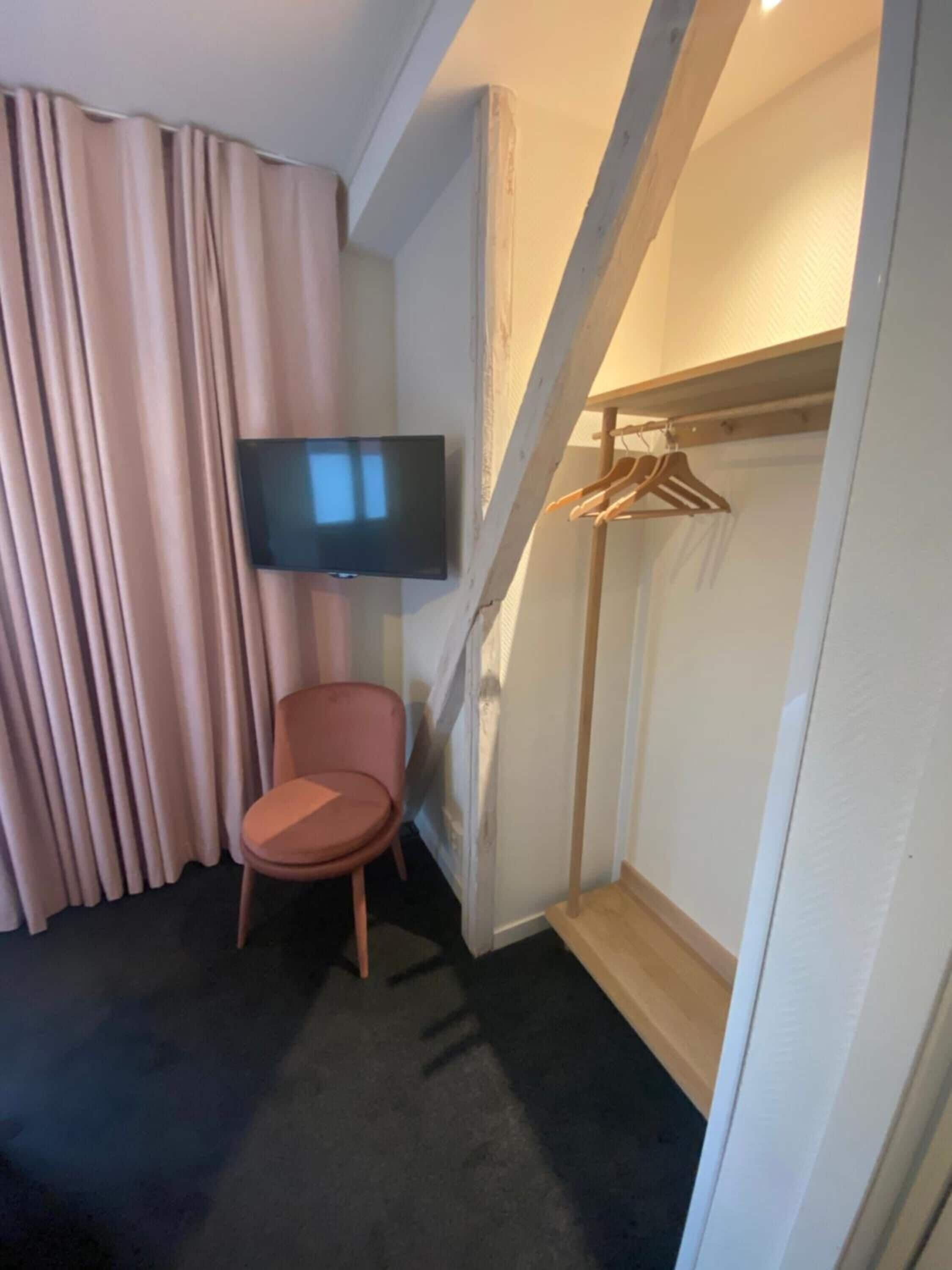 A hotel room with a flat-screen TV mounted on the wall, a chair, a coat rack, and a window with curtains.