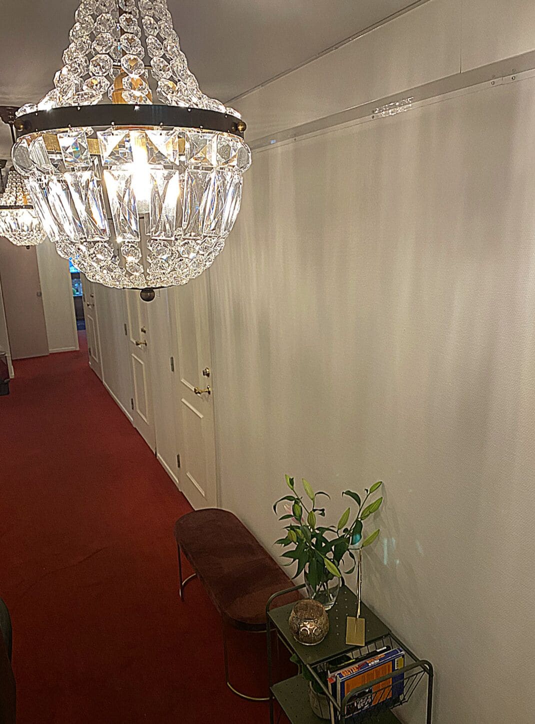 Un pasillo con candelabro, un banco, una mesita con planta y una caja, y una puerta.
