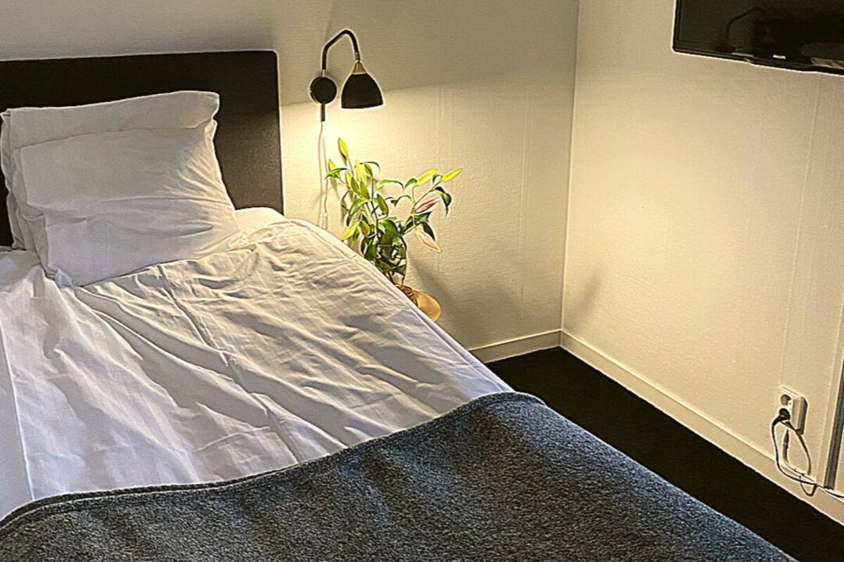 A neatly made bed with white linens and a dark gray throw blanket. There is a black lamp on a bedside table with a plant beside it. A flat-screen TV is mounted on the wall to the right of the bed.