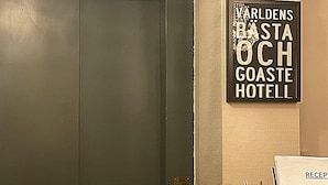 A hotel reception area with a closed elevator door, a sign for 'Världens Bästa Och Goaste Hotel', and a newspaper on the counter. - Hotell Värend (Växjö)