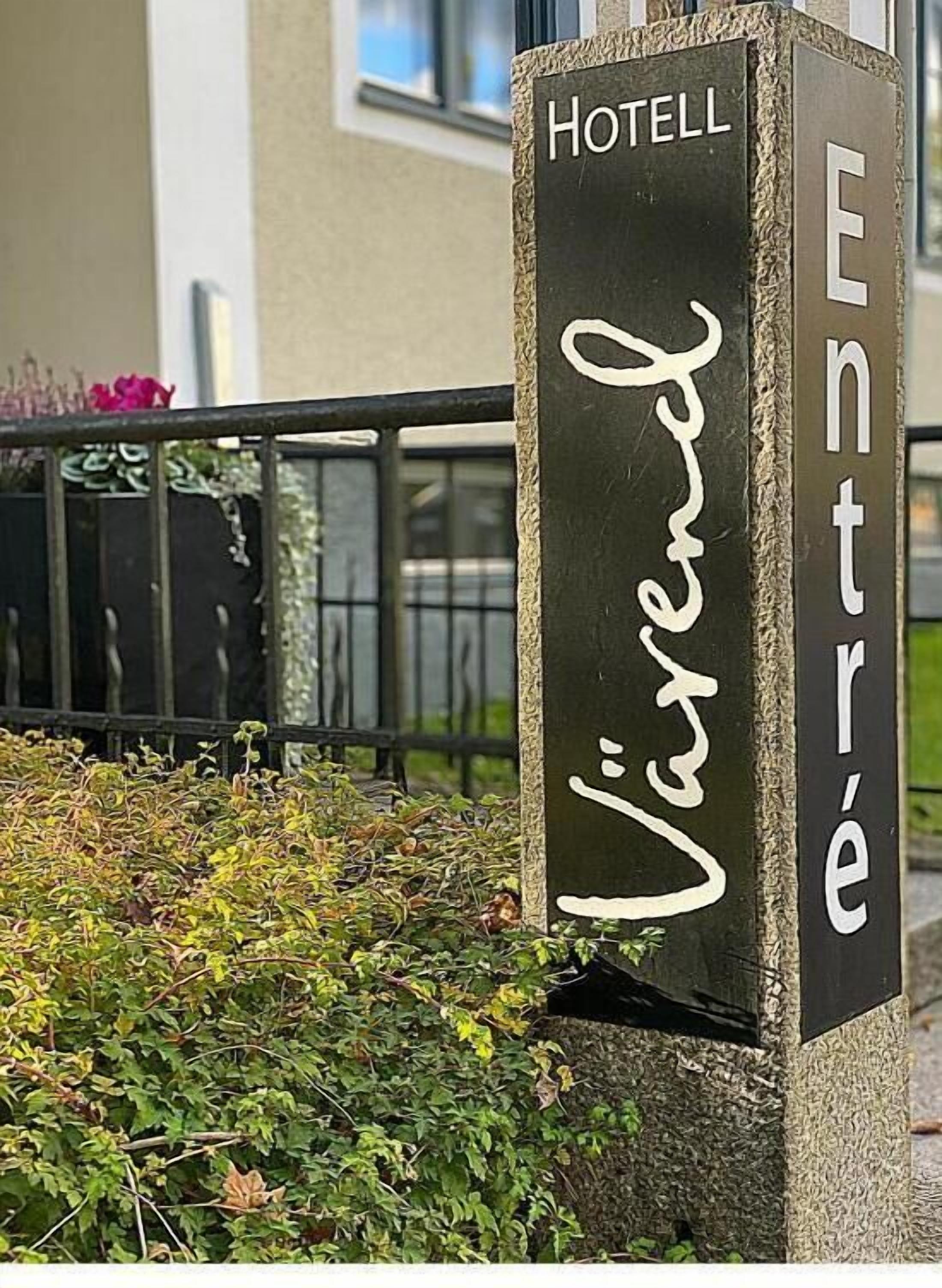 Ein Hotel Schild mit dem Schriftzug "HOTEL Entrelé" in weißen Buchstaben vor dunklem Hintergrund, umgeben von Grünpflanzen und einem Metallzaun.