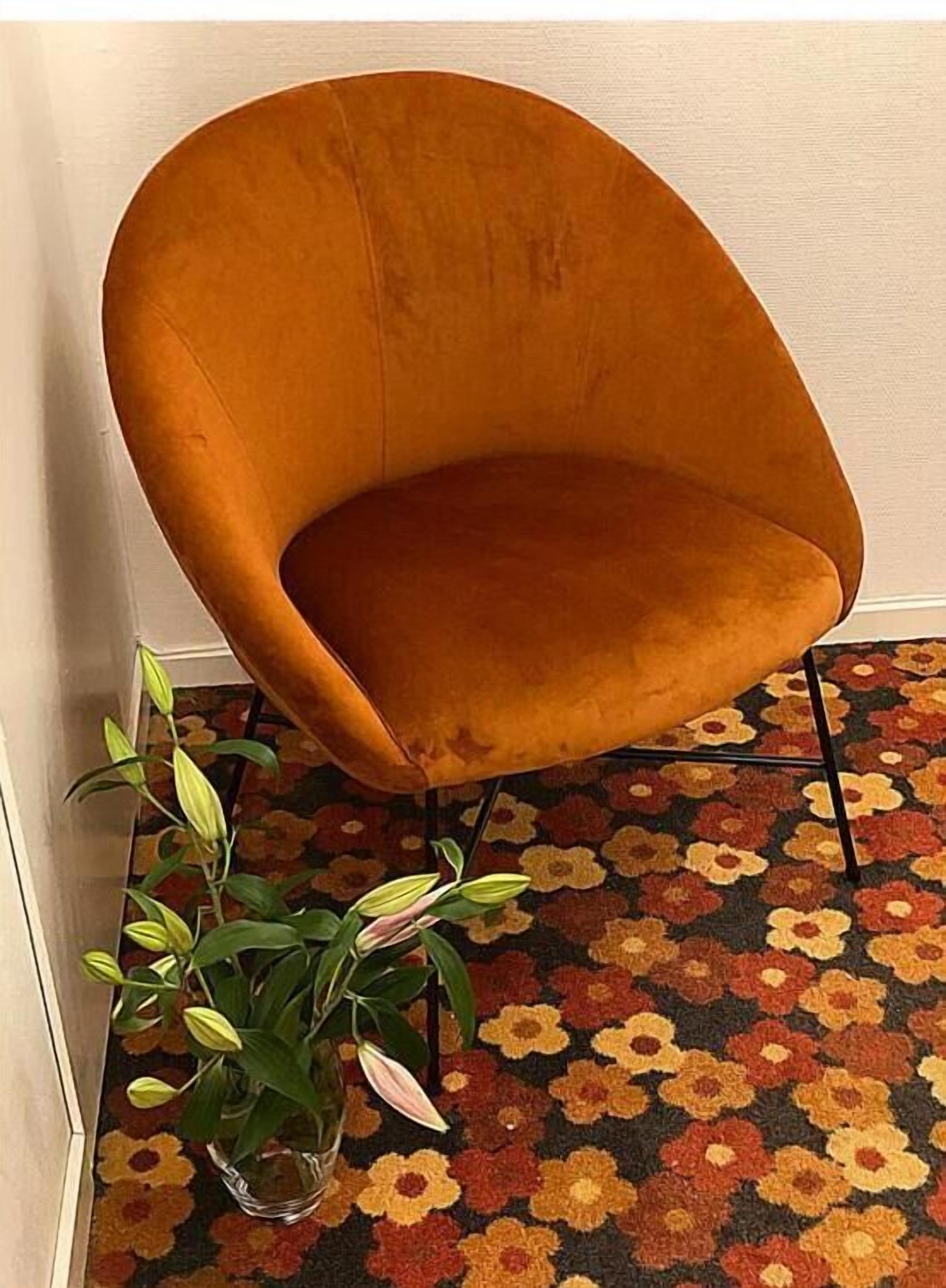 Une pièce d’angle avec un seul fauteuil orange, une plante en pot et un tapis à motifs floraux.