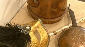 Une théière en cuivre dotée d’une anse et d’un bec verseur, un masque décoratif et un objet métallique rond posé sur une surface en bois.